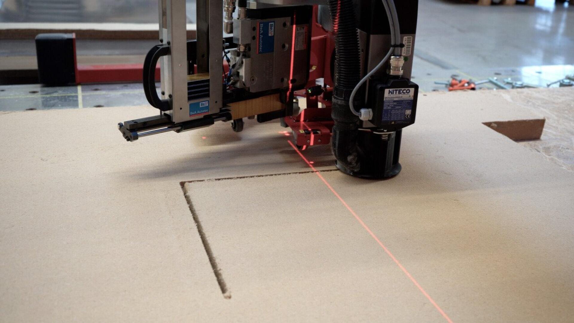 A close-up of a CNC router machine cutting a rectangular shape into a sheet of wood, with a red laser guide marking the path on the material.