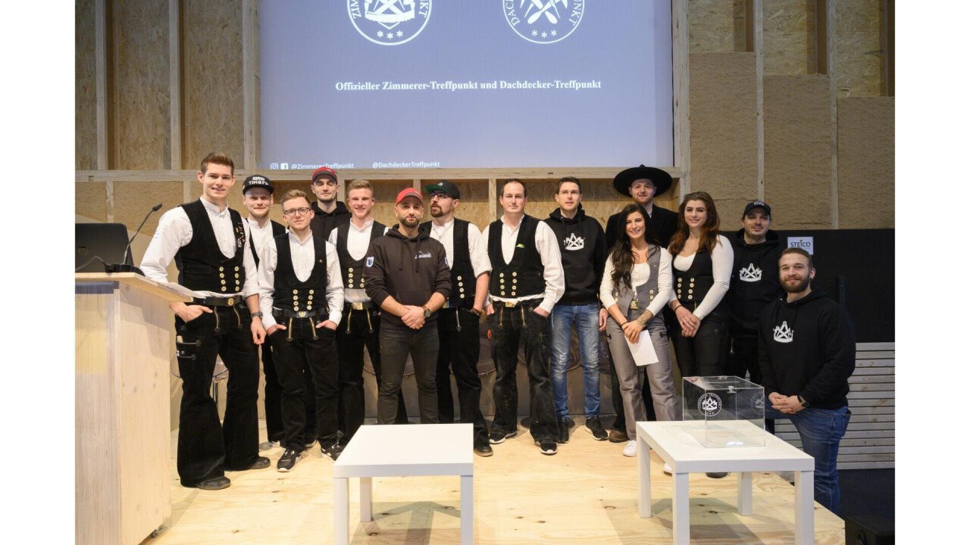 A group of people, some in traditional carpenter attire and others in casual clothing, stand together smiling on a stage with a logo and text projected on the wall behind them.