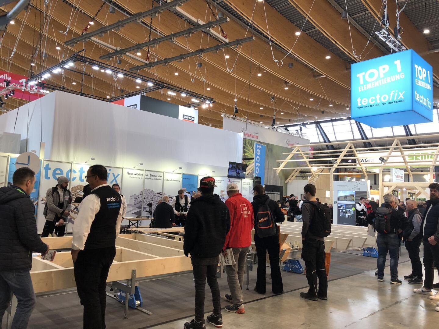 Menschen gehen durch eine belebte überdachte Messe mit Holzrahmen, Bannern und Ständen. Mehrere Personen tragen Markenkleidung, und über dem Ausstellungsbereich prangt ein großes beleuchtetes Schild mit der Aufschrift tectofix.