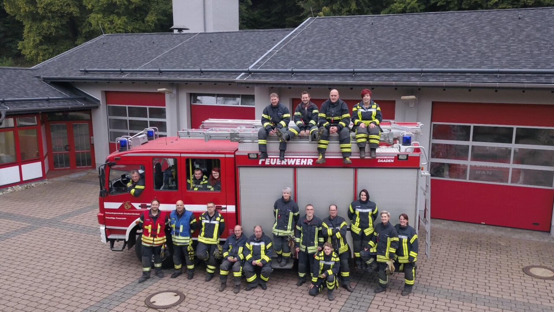 Eine Gruppe von Feuerwehrleuten in Uniform posiert vor einem roten Feuerwehrauto, das vor einer Feuerwache geparkt ist. Einige stehen auf dem Boden, während andere auf dem Fahrzeug sitzen. Die Feuerwache hat rote Türen und einen gefliesten Innenhof.