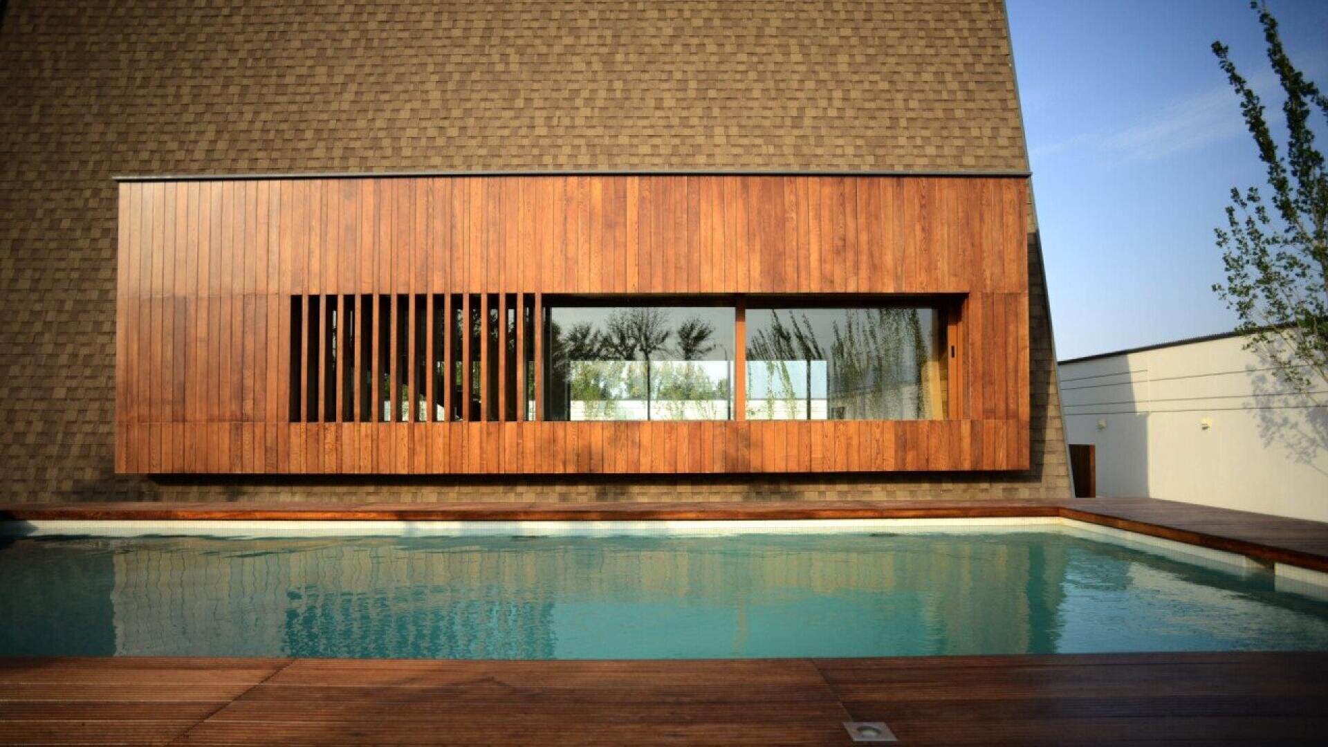 Ein modernes Gebäude mit einem großen, holzgetäfelten Fenster blickt auf einen klaren Swimmingpool. Die Struktur besteht aus einer braunen Schindelwand und einem Holzdeck, in dessen Glas sich der blaue Himmel und die Bäume spiegeln.