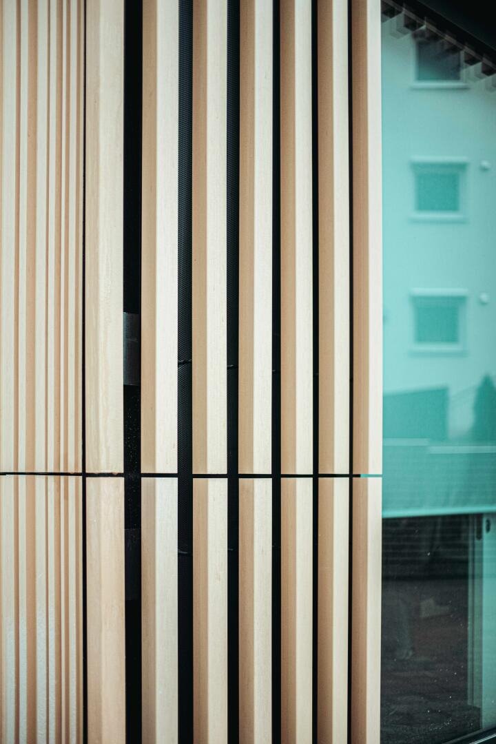 Nahaufnahme von vertikalen Holzlatten, die Teil einer modernen architektonischen Fassade sind, wobei sich in einer Glasfläche auf der rechten Seite ein Gebäude spiegelt.
