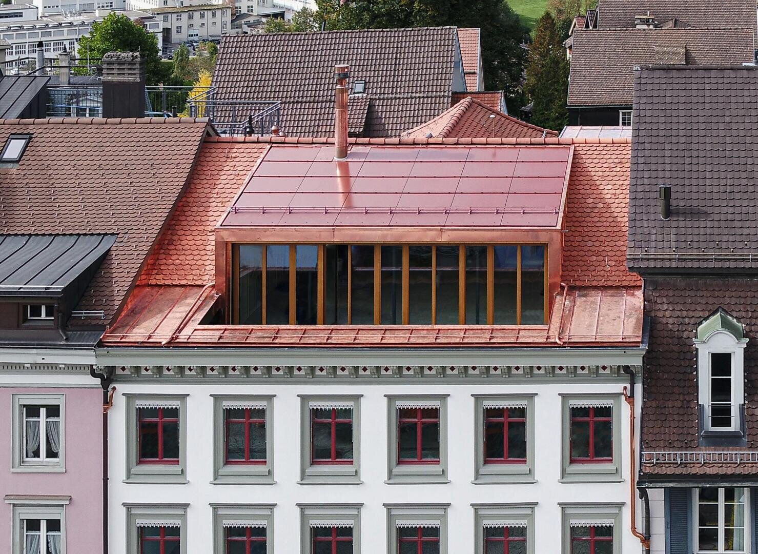 Ein renoviertes Gebäude mit rotem Ziegeldach und großen modernen Oberlichtfenstern, umgeben von traditionellen Gebäuden in einem städtischen Viertel. Die Fassade ist mit grünen Zierleisten und rot gerahmten Fenstern versehen.