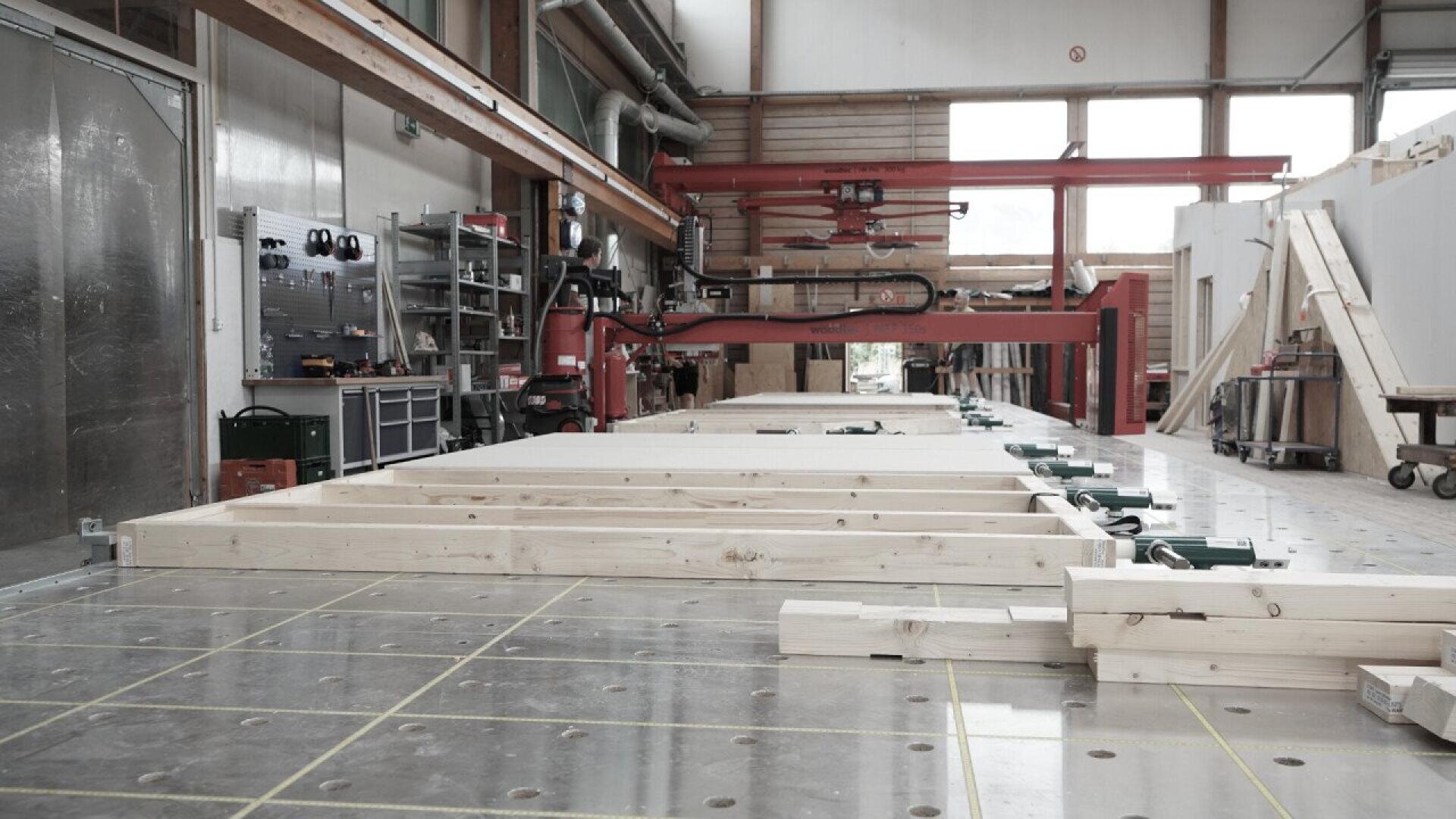 A spacious woodworking workshop with wooden beams laid out on a large worktable, surrounded by various tools, equipment, shelves, and a red overhead crane in the background.