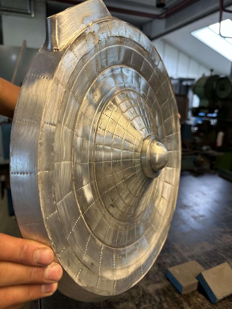 Eine Person hält in einer Werkstatt ein glänzendes, metallisches, rundes Objekt mit eingravierten konzentrischen Ringen und einem spitzen Mittelpunkt, das einem detaillierten Raumschiff- oder Satellitenmodell ähnelt.