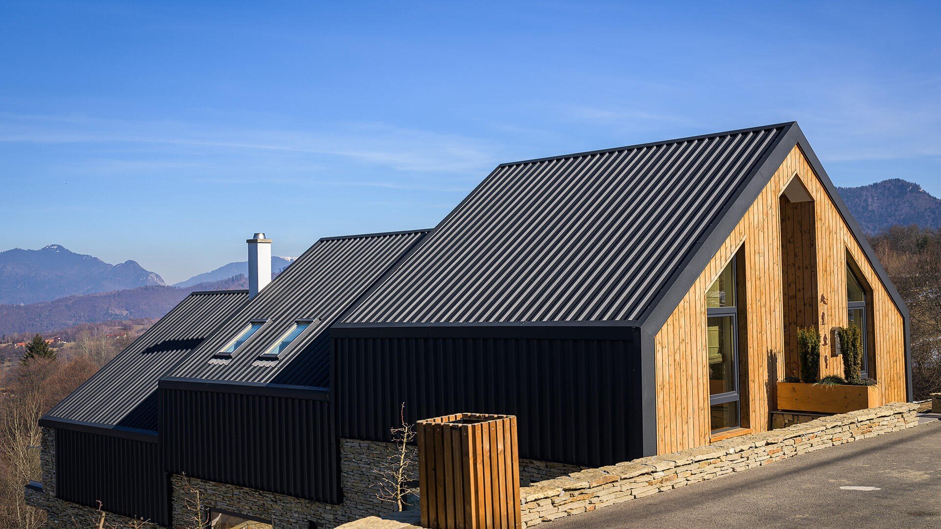 Ein modernes Haus mit schwarzen Metalldächern und Holzwänden steht auf einer Steinterrasse in einer bergigen Landschaft unter blauem Himmel.