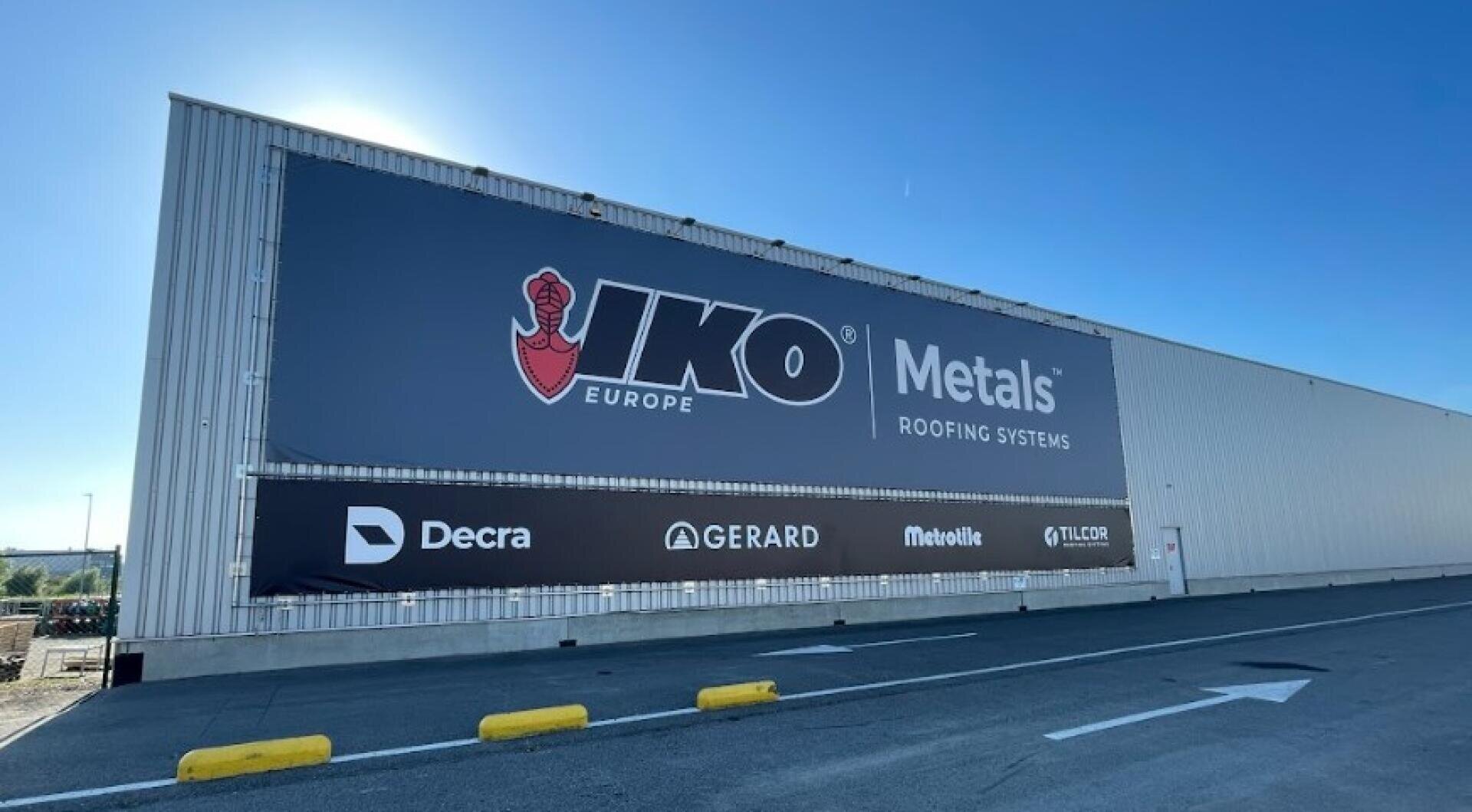 Ein großes Metallgebäude mit einem auffälligen Schild für IKO Metals Roofing Systems und Logos für die Marken Decra, Gerard, Metrotile und Tilcor steht unter einem strahlend blauen Himmel.