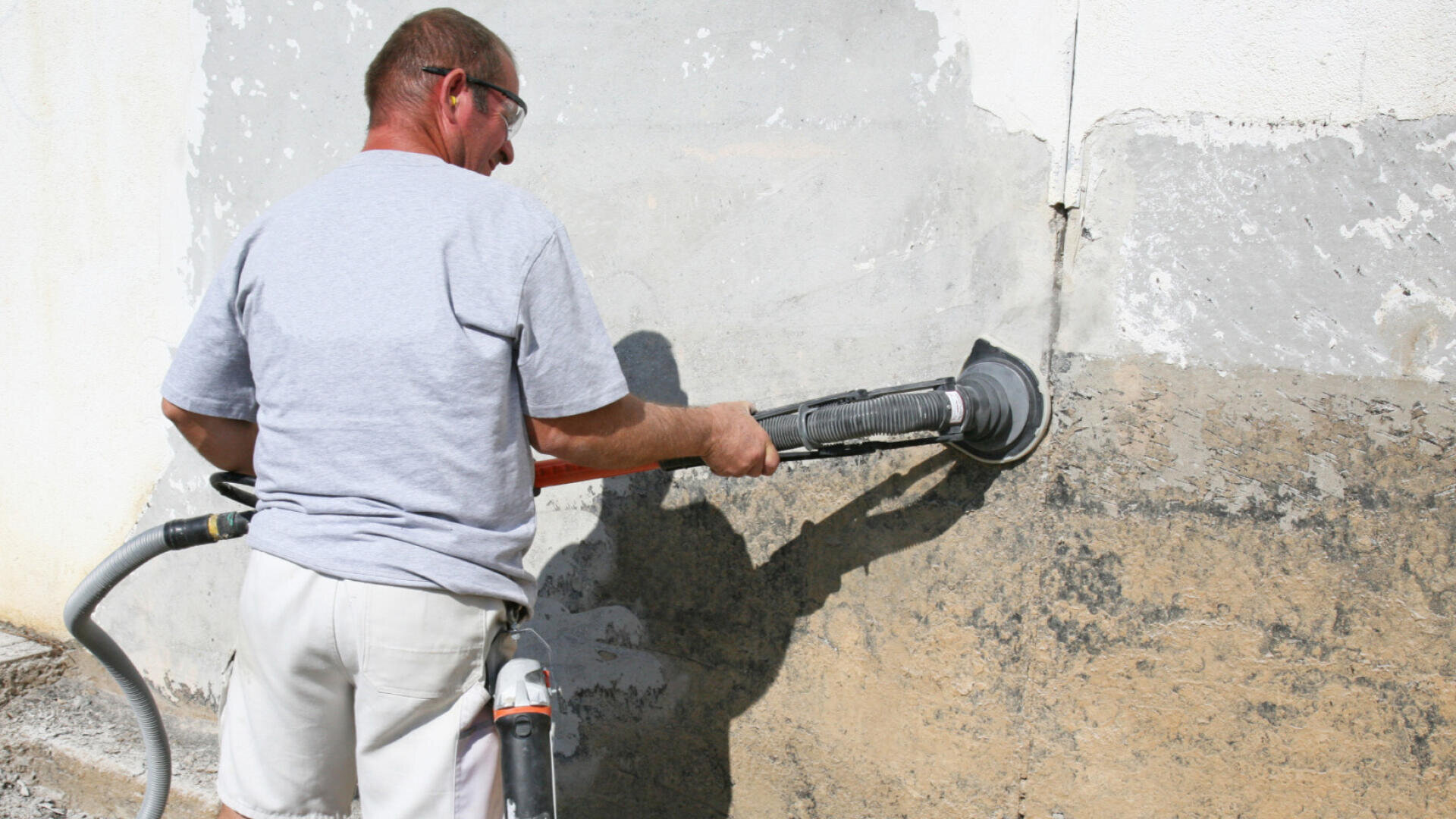 A man wearing safety goggles is using an electric sander to remove old paint and plaster from an exterior wall. He is dressed in a gray T-shirt and light-colored shorts and has a vacuum cleaner attachment connected to the sander.
