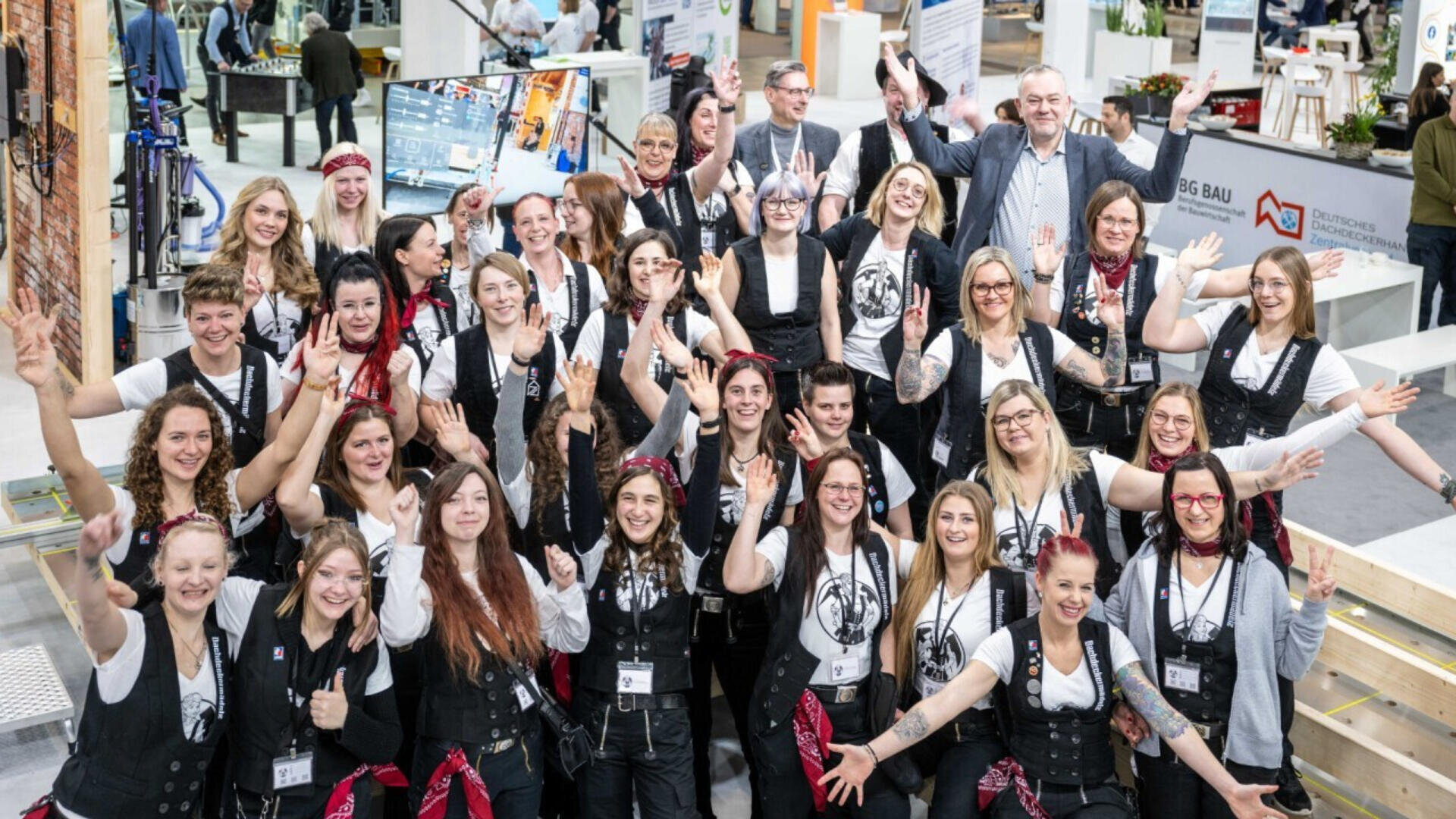 A large group of smiling people, mostly women, pose together indoors at an event, many wearing matching black vests, white shirts, and red bandanas, raising their arms and making peace signs.