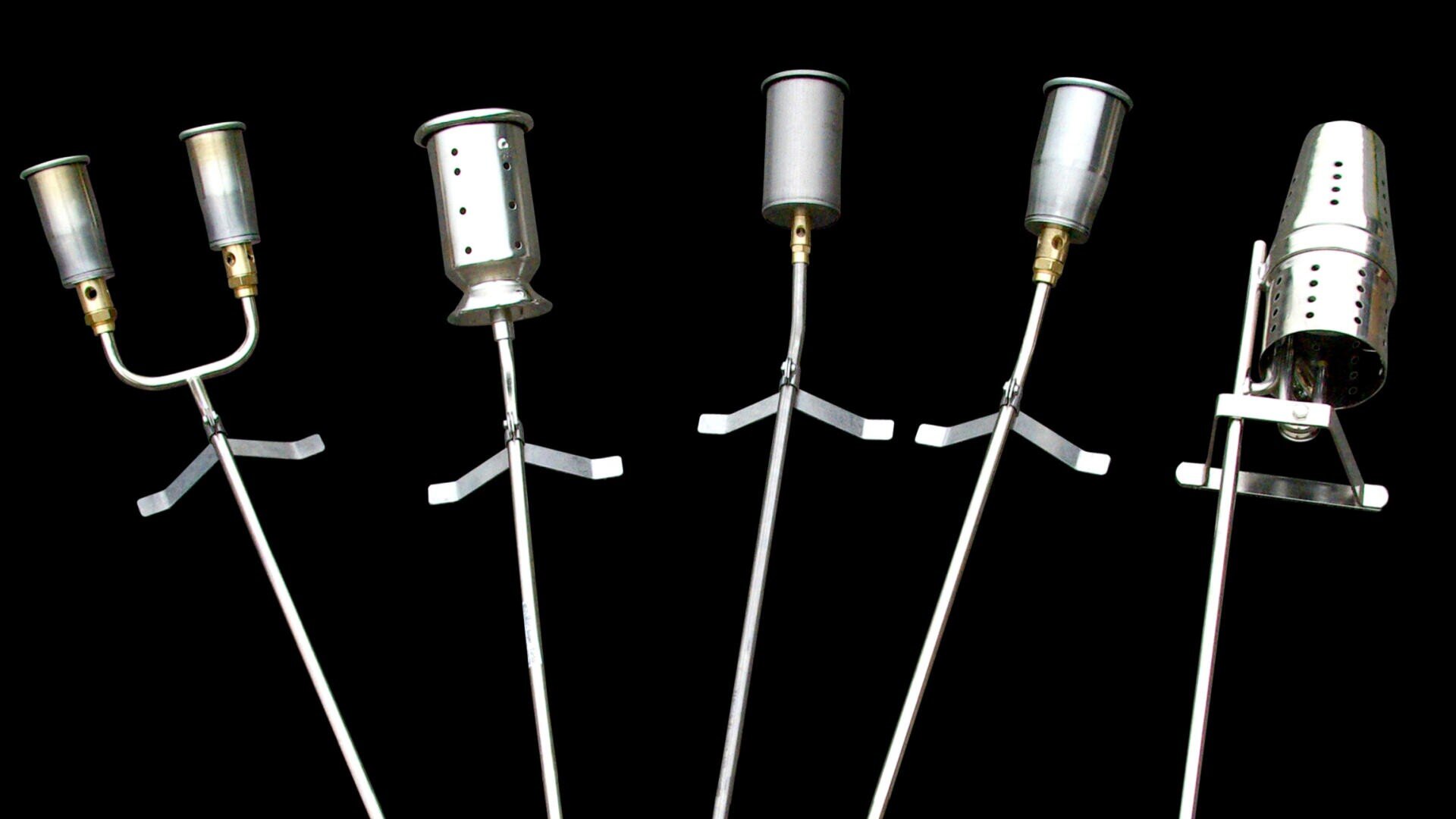 Five different metal weed burners with long handles and various nozzle designs are displayed upright against a black background.
