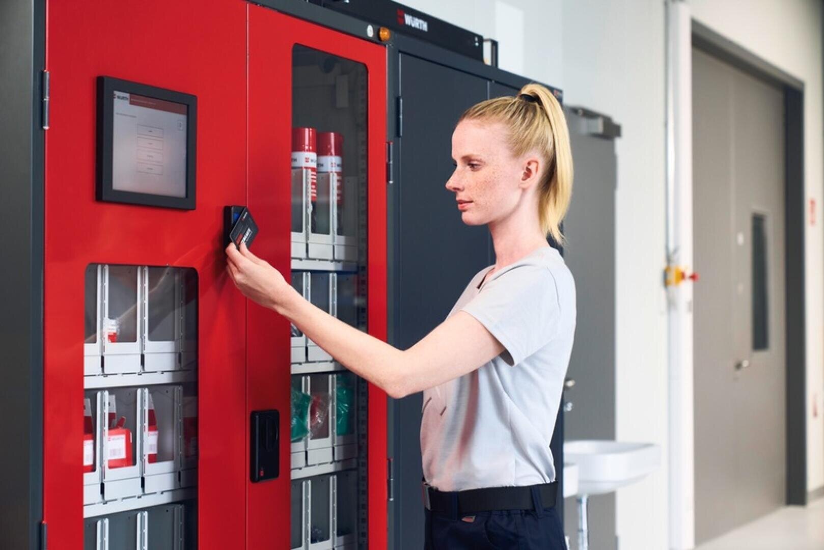 Eine Frau benutzt eine Schlüsselkarte, um einen roten Automaten mit transparenten Fächern, die verschiedene Artikel enthalten, in einem modernen, gut beleuchteten Innenraum zu öffnen.