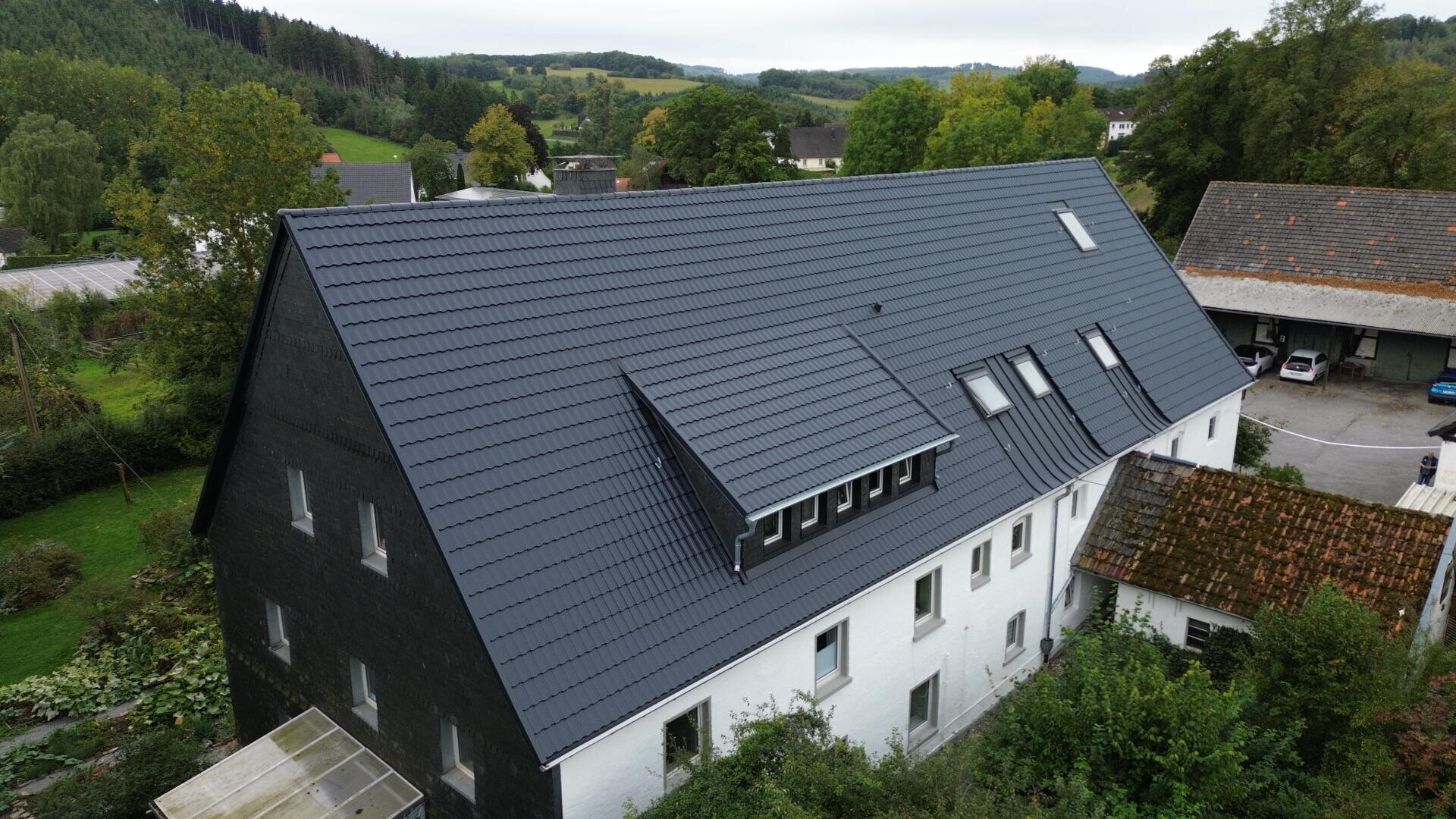 A large white building with a dark, sloped roof featuring multiple windows and skylights, surrounded by trees and greenery in a rural area with hills in the background.