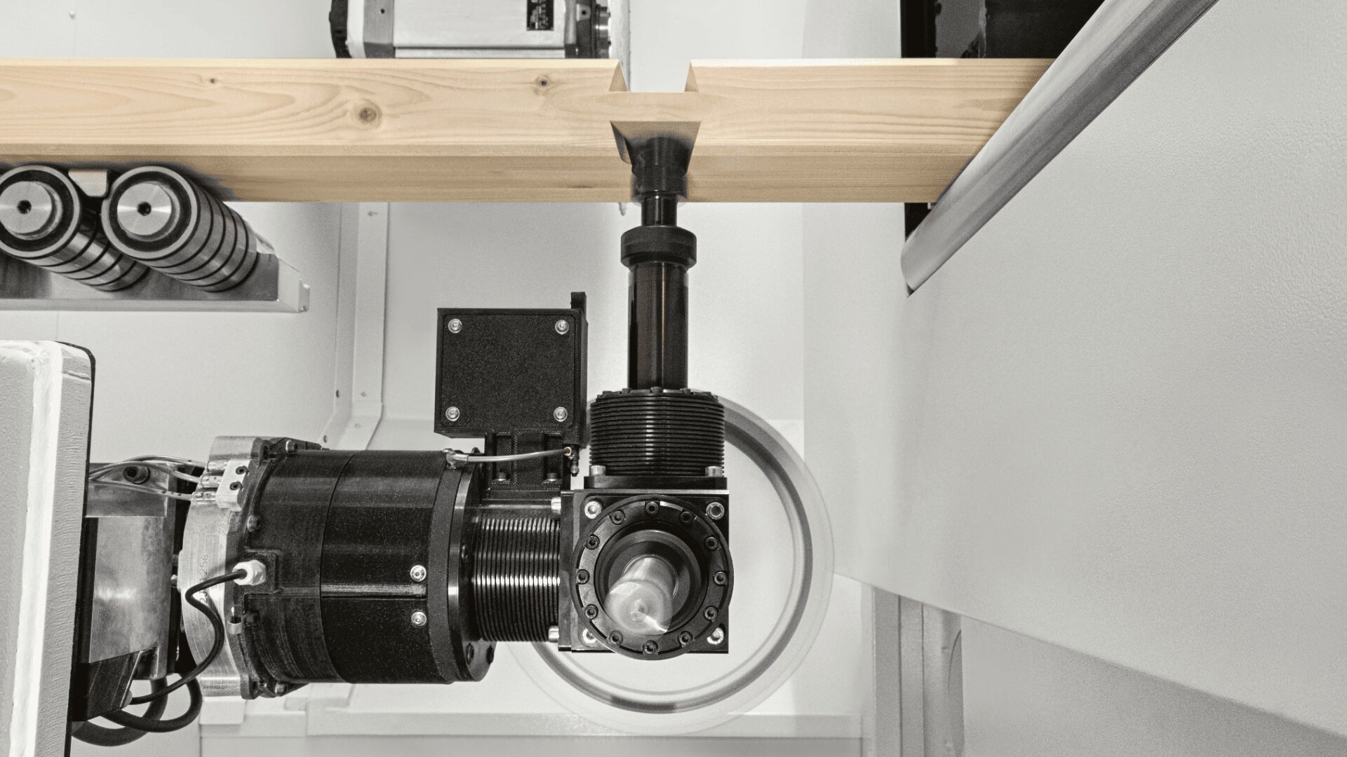 Nahaufnahme eines Roboterarms mit Schneidwerkzeug, der in einer Industrie- oder Fertigungsumgebung einen Holzbalken bearbeitet. Im Hintergrund sind Metallkomponenten und Maschinen zu sehen.