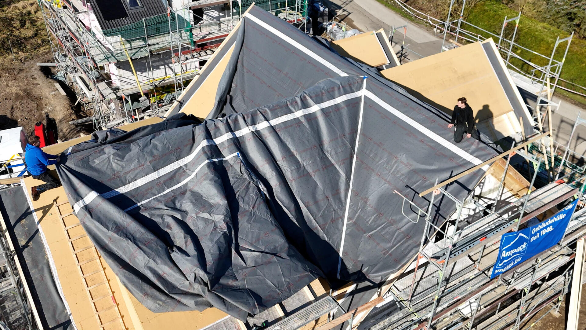 Aerial view of a building under construction with scaffolding. The roof is covered with a large gray tarp, and two people are standing on either side of the structure. Building materials and tools are visible.