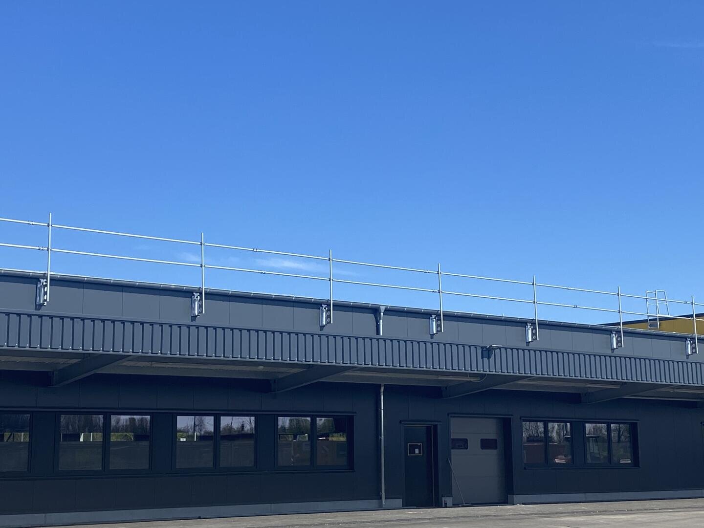 An industrial building with a flat roof, dark gray walls and several windows, with a safety railing along the edge of the roof, under a clear blue sky.
