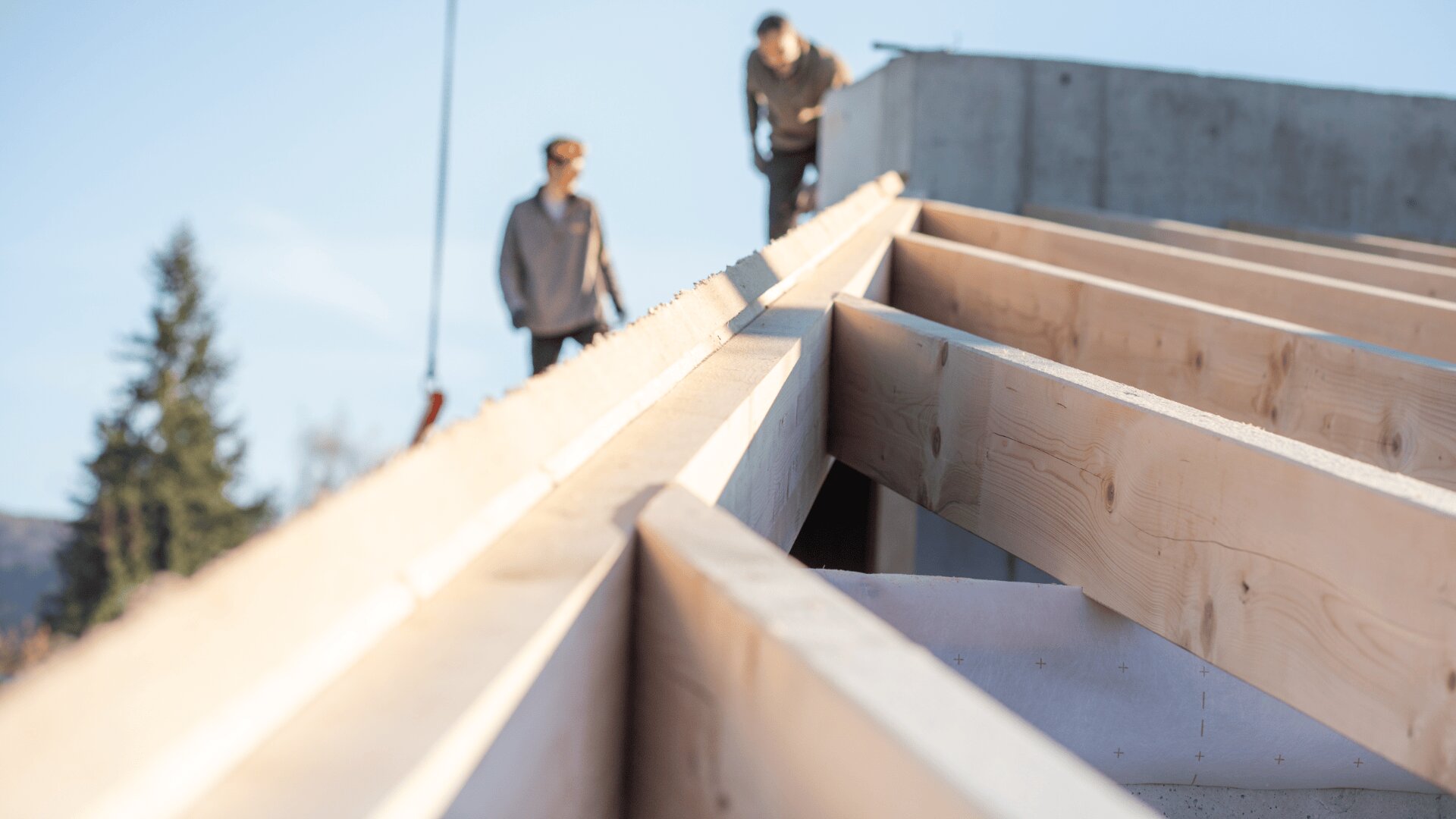 Nahaufnahme von im Bau befindlichen hölzernen Dachbalken. Im Hintergrund stehen zwei unscharfe Personen an einem sonnigen Tag auf einer teilweise fertiggestellten Struktur.