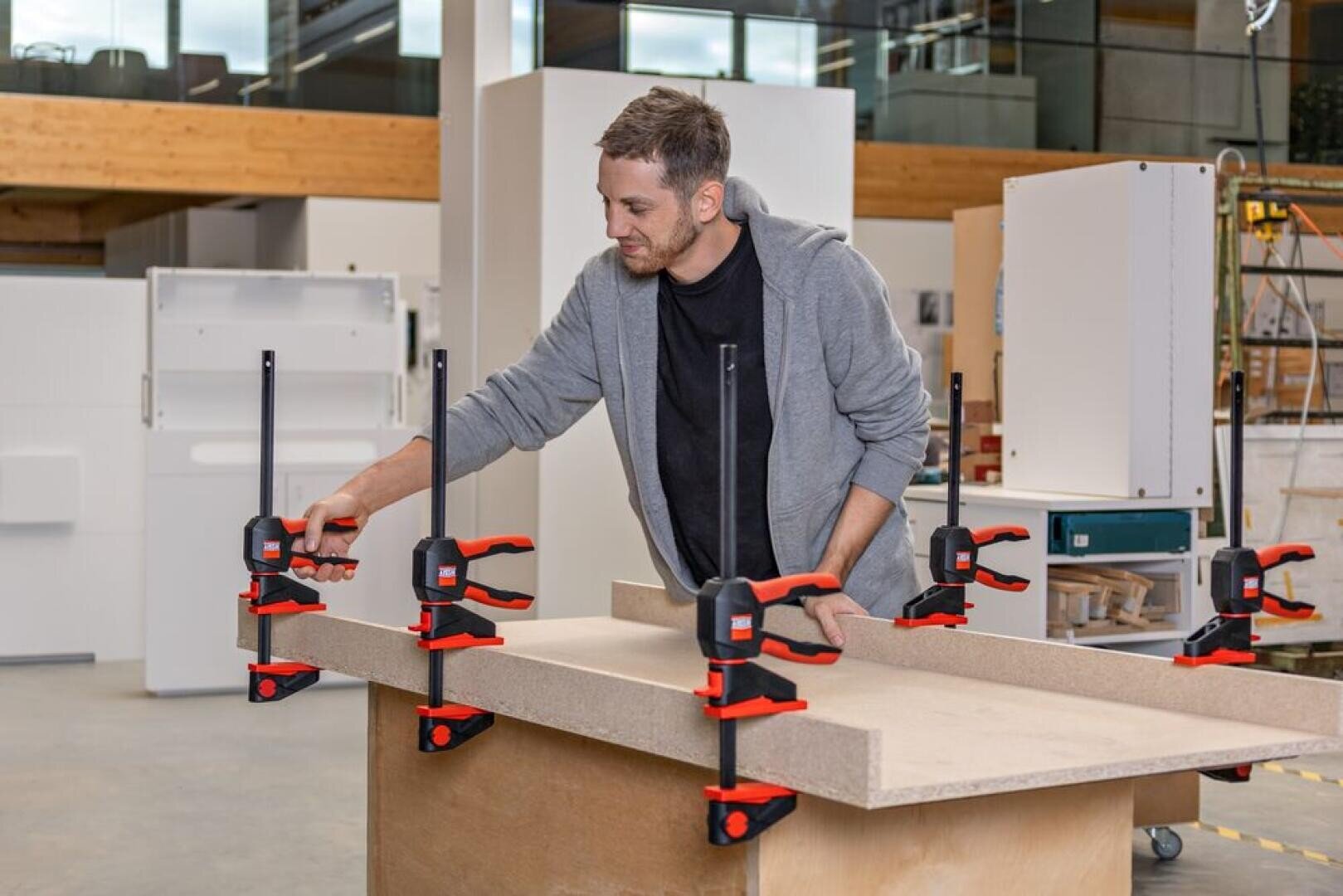 A man in a workshop uses several red and black clamps to secure two large wooden boards together on a workbench, surrounded by cabinets and tools.