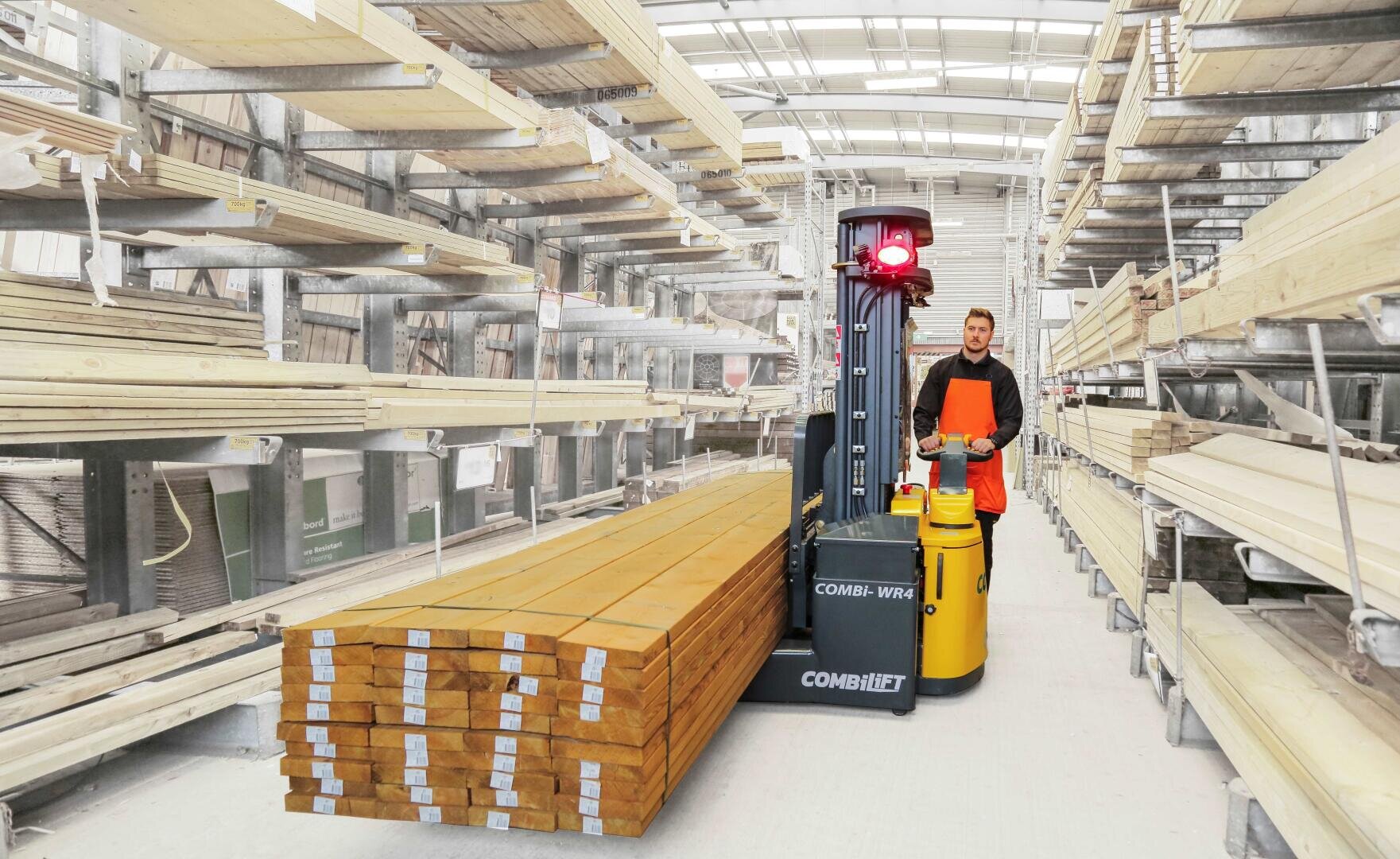 Ein Arbeiter in einer orangefarbenen Sicherheitsweste bedient einen Gabelstapler und transportiert einen Stapel langer Holzbretter durch ein Holzlager mit Regalen voller Holzplatten.