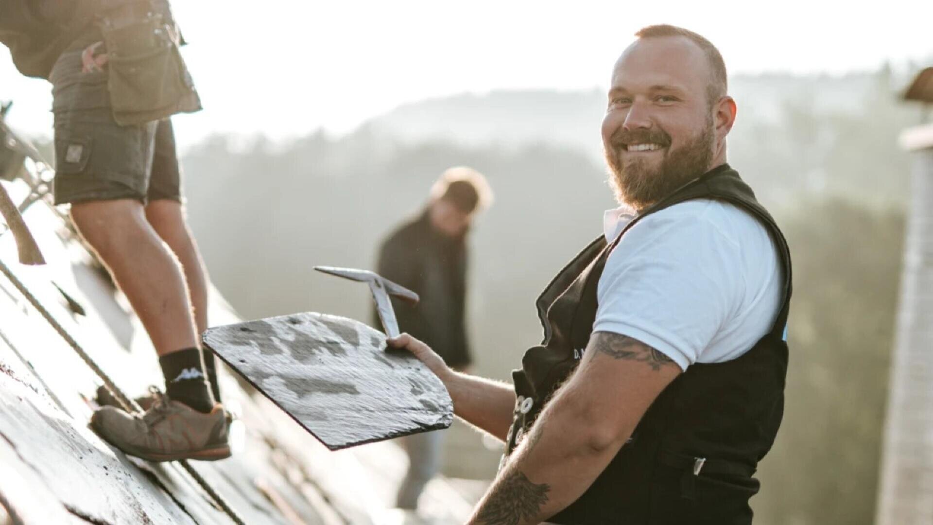A smiling man holding a roofing tool sits on a sloped roof, wearing a black vest and white shirt. Another person’s legs are visible beside him. The background is blurred, suggesting an outdoor setting.