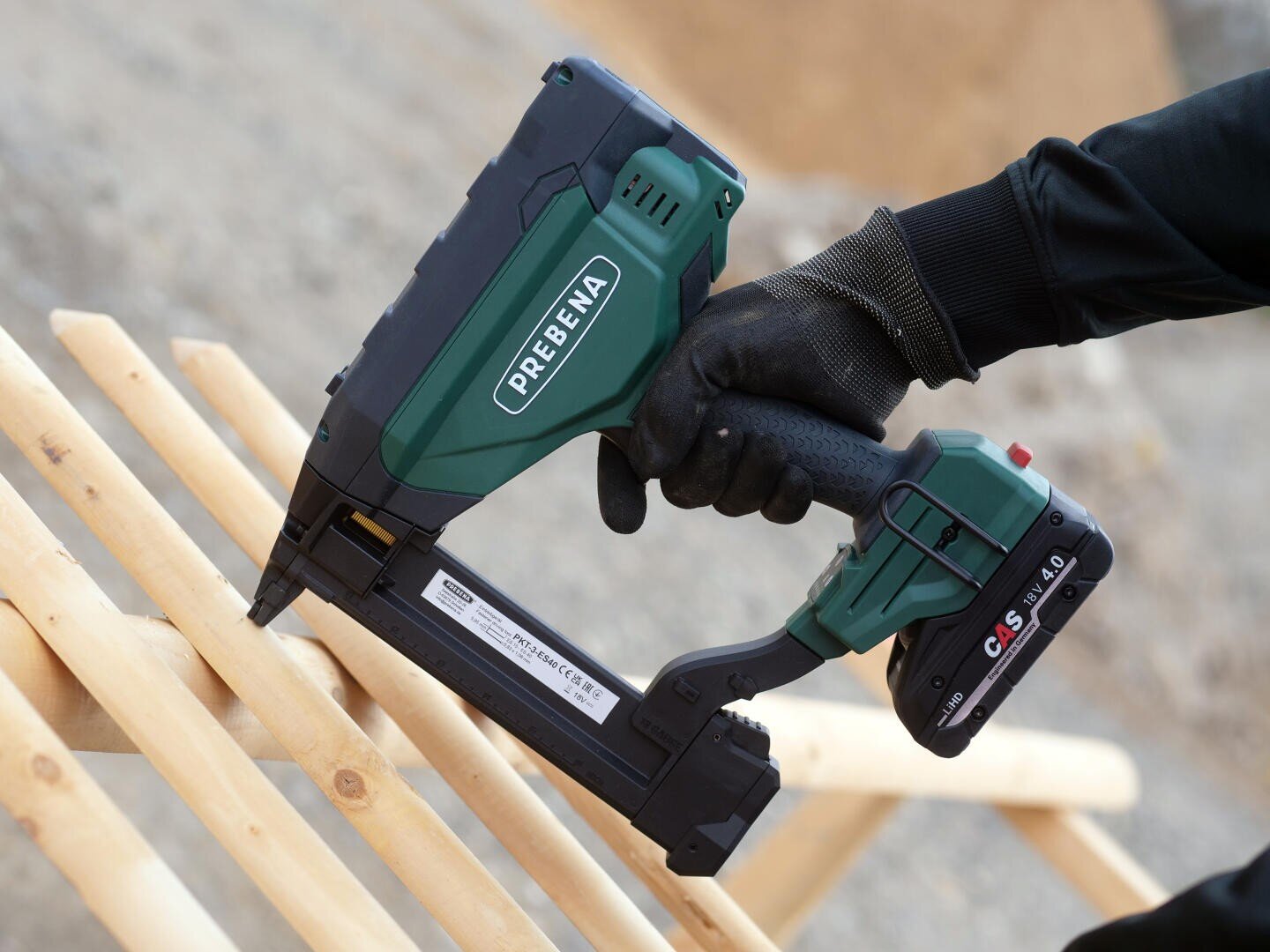 Eine Person mit schwarzem Handschuh benutzt eine grün-schwarze kabellose PREBENA-Nagelpistole, um Holzbalken im Freien zu befestigen.