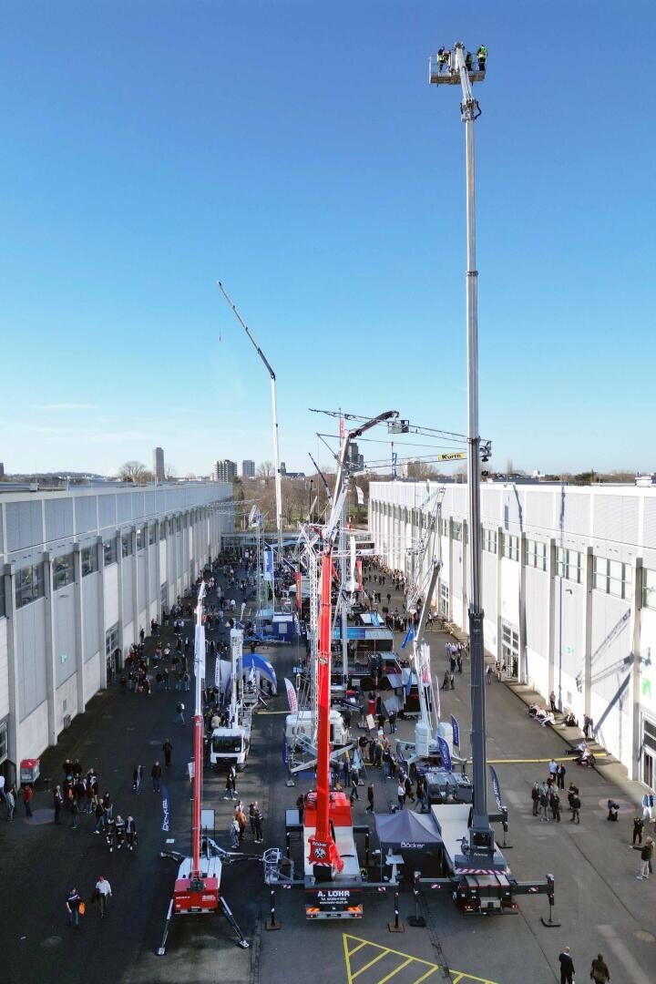 In einer Freiluftausstellung werden zwischen zwei Gebäuden große Baukräne und Maschinen ausgestellt, um die die Besucher herumgehen und die Geräte unter blauem Himmel betrachten können.