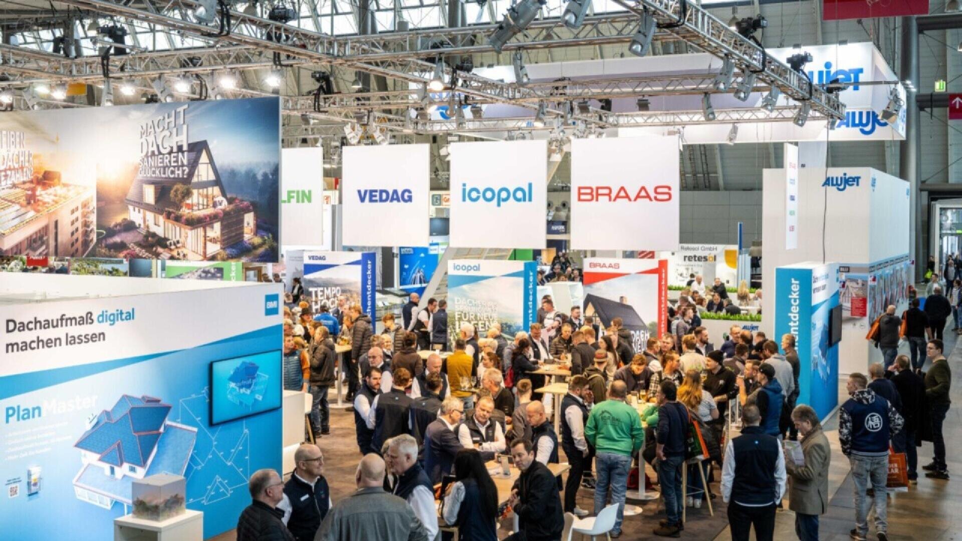 A large crowd of people exploring various company booths at a busy indoor trade show or exhibition, with colorful displays, banners, and informational signs overhead.
