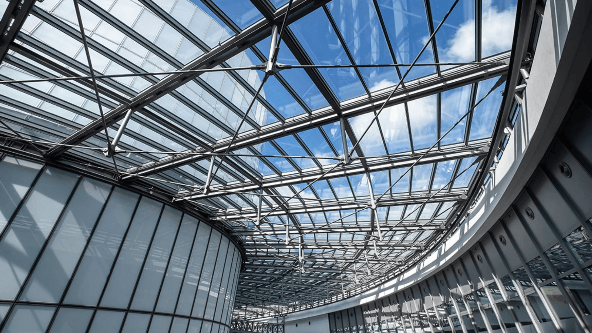 Gebogene Dachkonstruktion aus Glas und Stahl mit gitterartigem Muster, die natürliches Licht in das Innere des modernen Gebäudes lässt und den Blick auf einen blauen Himmel mit einigen Wolken freigibt.