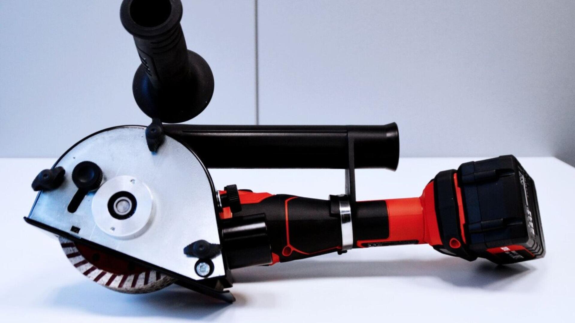 A red and black cordless angle grinder with a side handle is placed on a white surface against a plain, light-colored background.