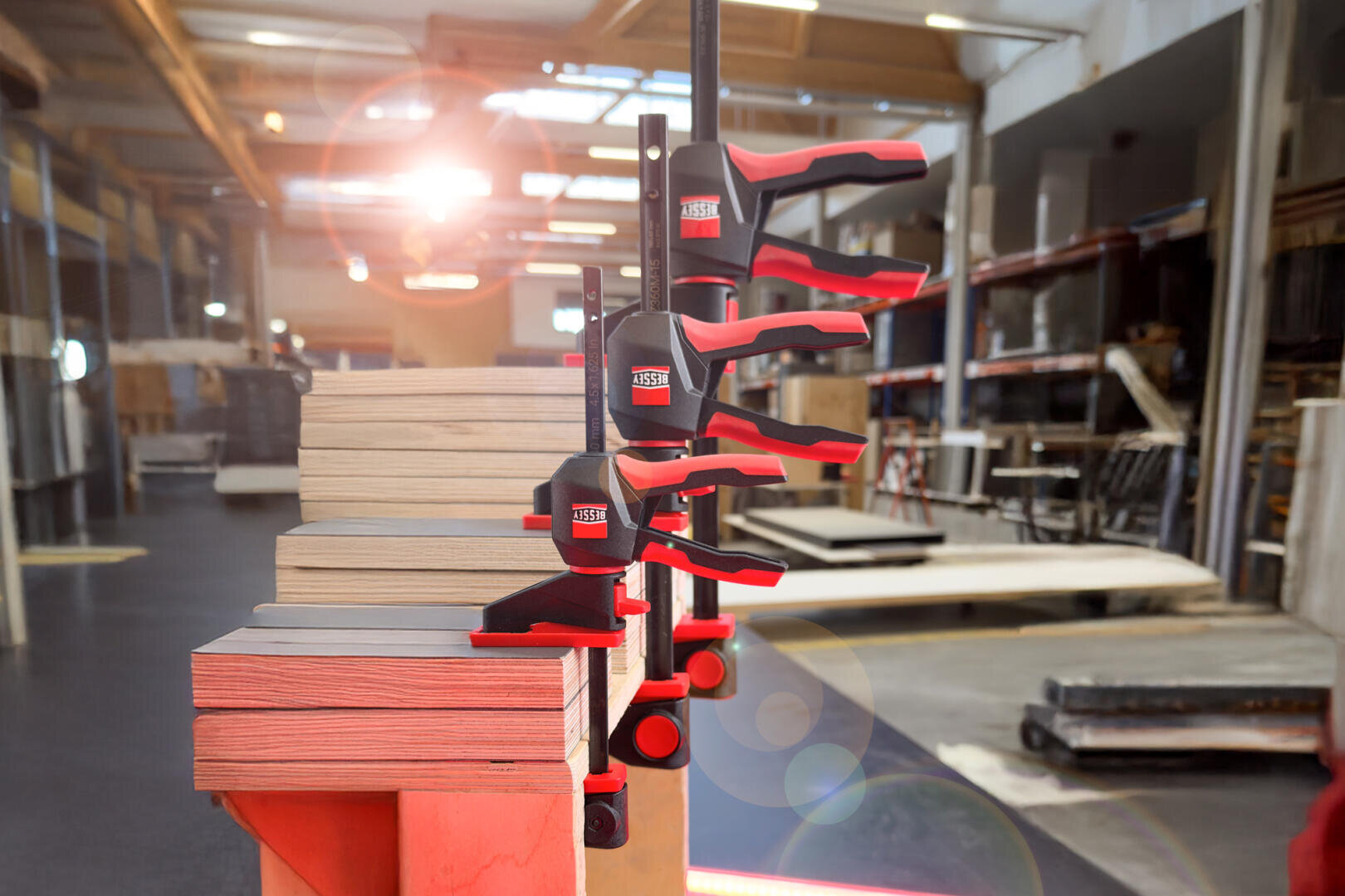 Four red and black clamps hold stacked wood pieces together in a bright, modern workshop with shelves and tools visible in the background. Sunlight streams in from the upper left corner.