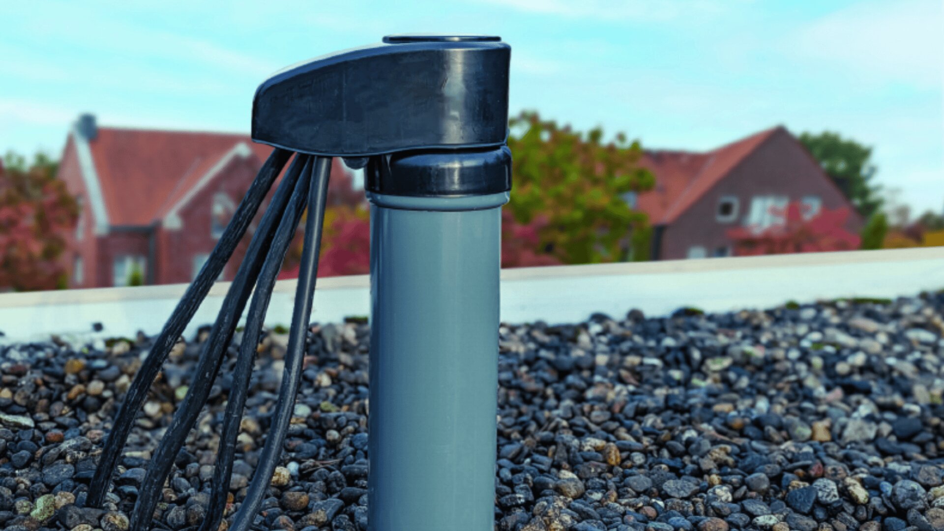 Nahaufnahme eines grauen Dachentlüftungsrohrs mit daran befestigten schwarzen Kabeln, umgeben von feinem Kies. Im Hintergrund sind verschwommene Häuser und Bäume unter blauem Himmel zu sehen.