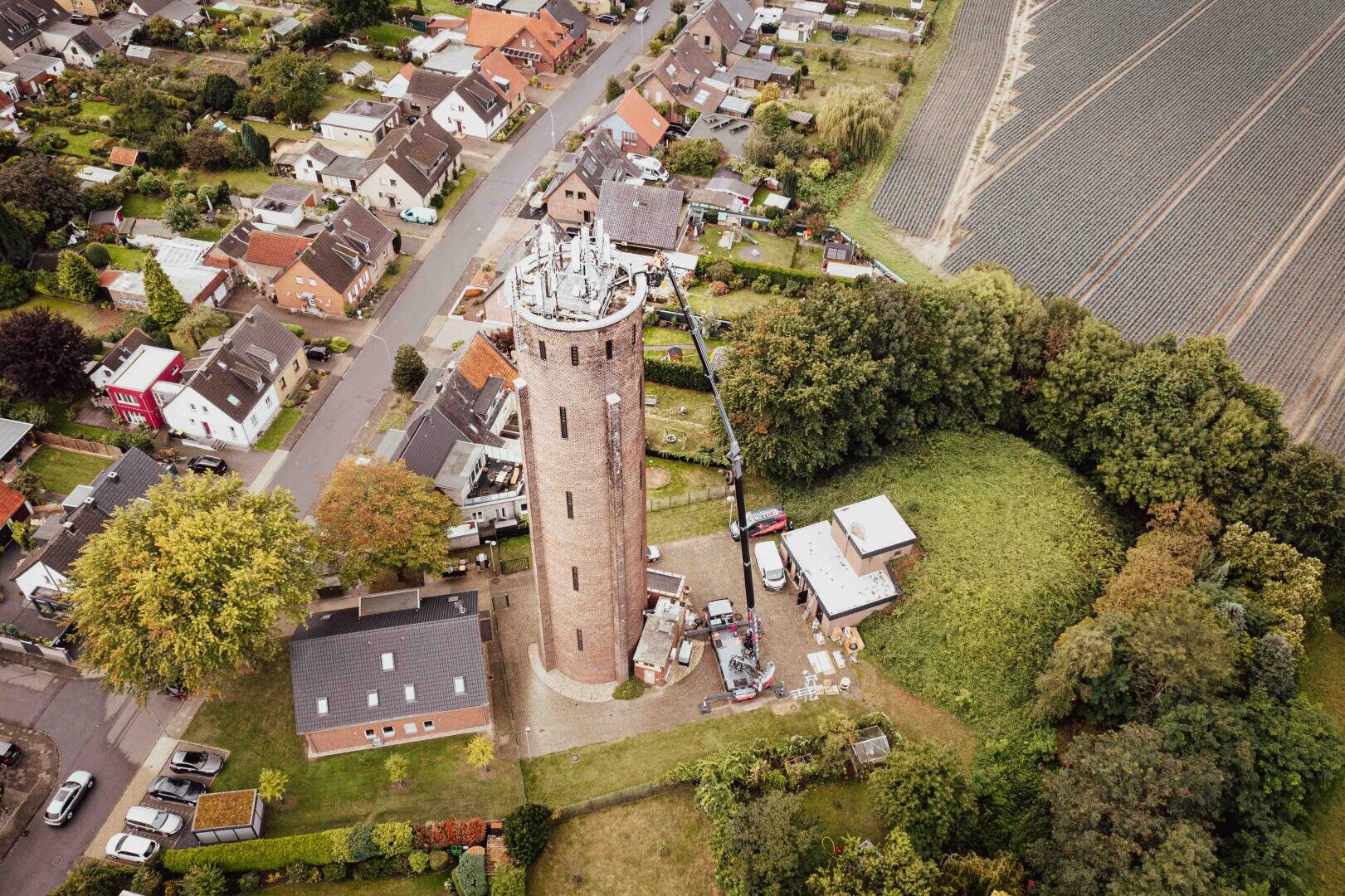 Aerial view of a tall, round brick water tower surrounded by trees and houses, with farmland on the right and roads leading through a residential neighborhood.