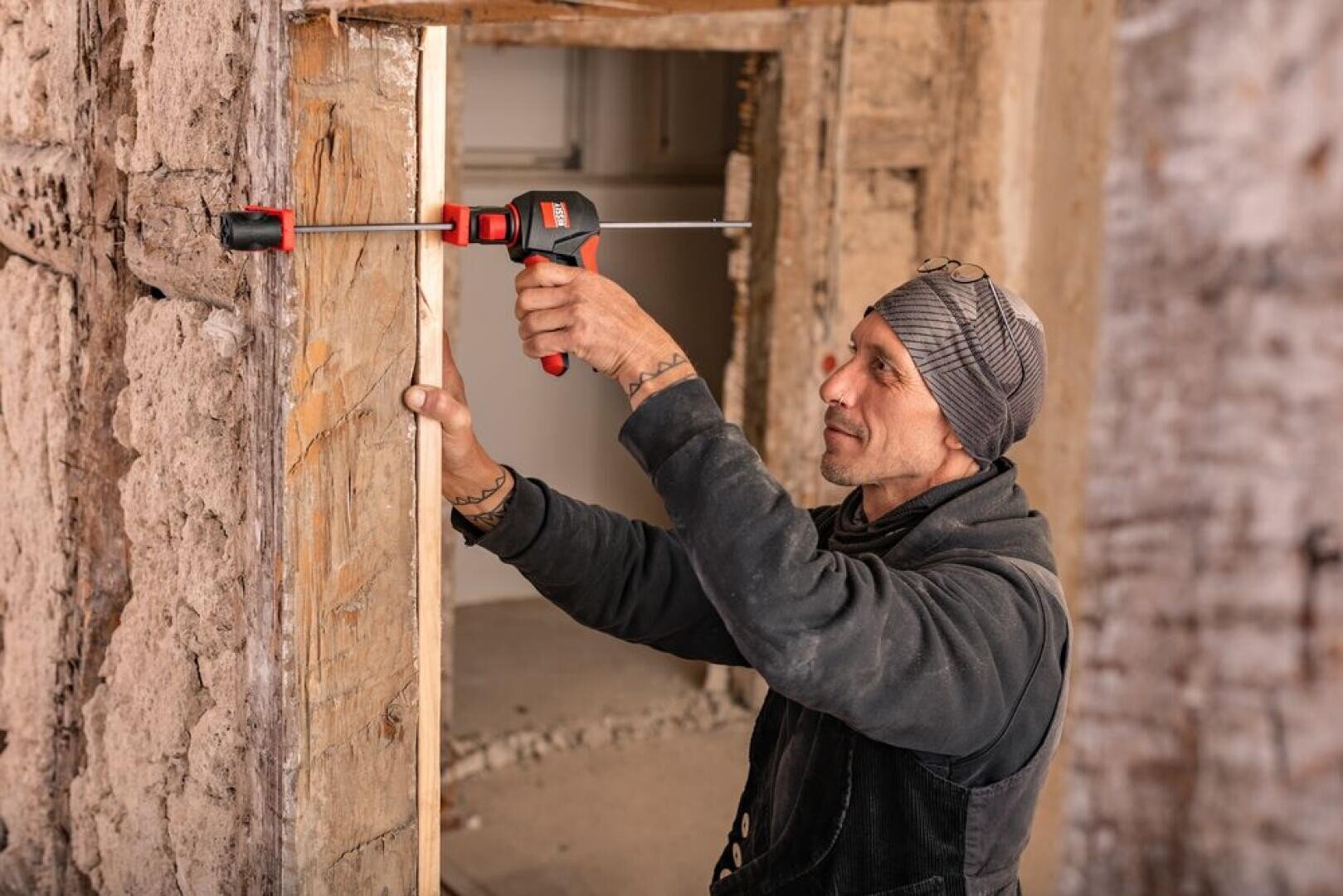 A construction worker in a cap uses a clamp to attach a wooden board to a rough door frame in a building under renovation.