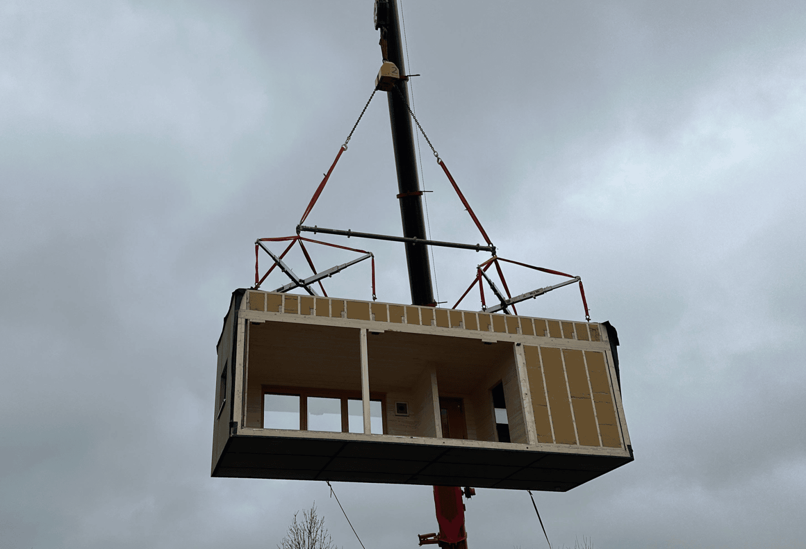 A crane lifts a large prefabricated building module with visible windows and door openings against a cloudy sky.