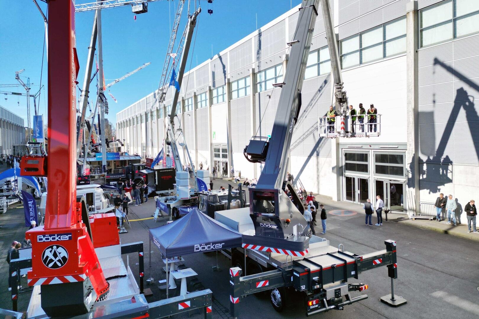 Ausstellung von Baumaschinen im Freien mit mehreren großen Kränen und Hebemaschinen, mit Menschen, die auf einer erhöhten Plattform in der Nähe eines modernen Industriegebäudes unter strahlend blauem Himmel fahren und beobachten.