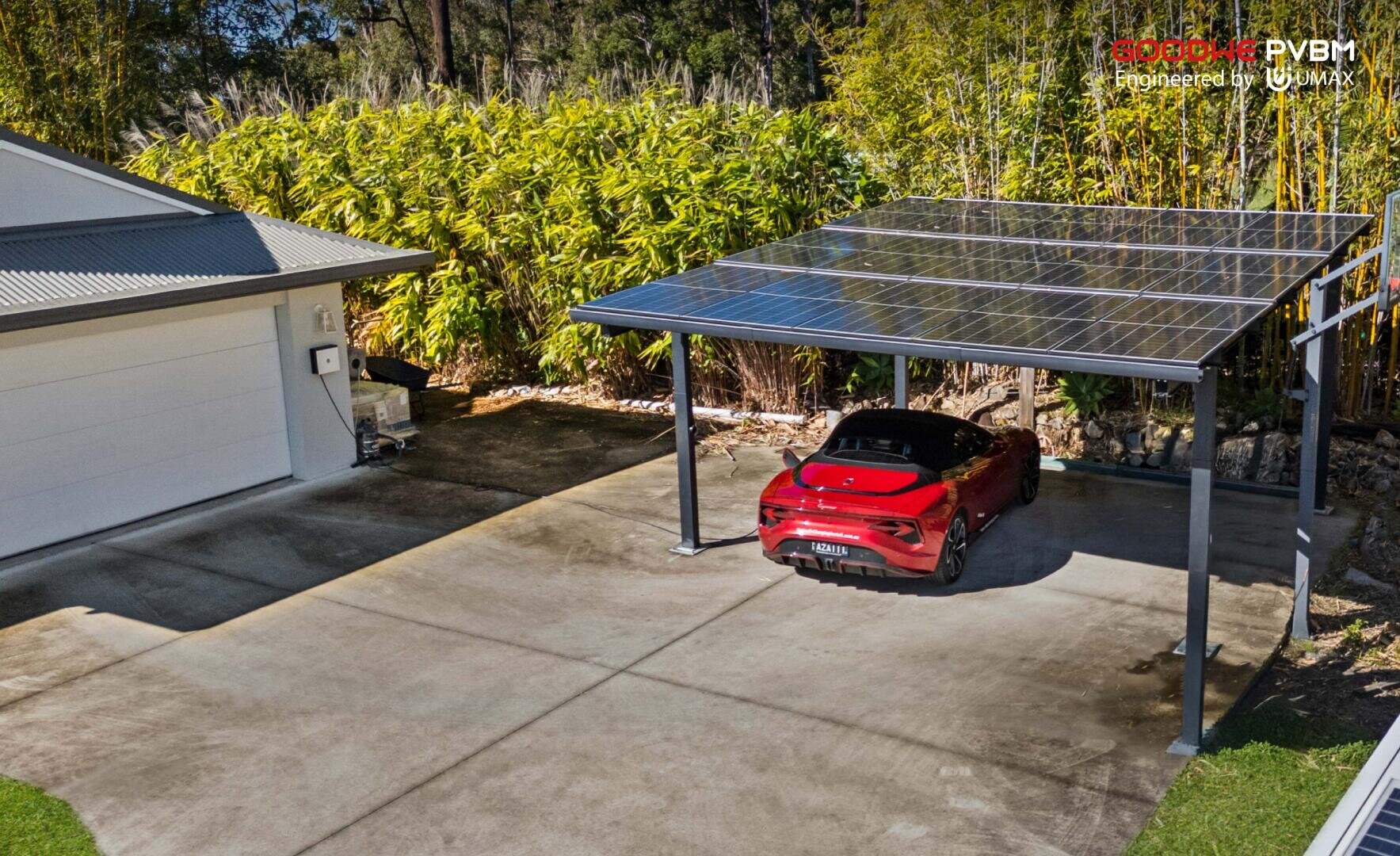 A red sports car is parked under a modern carport with a transparent solar panel roof next to a house surrounded by tall green bamboo and a concrete driveway.