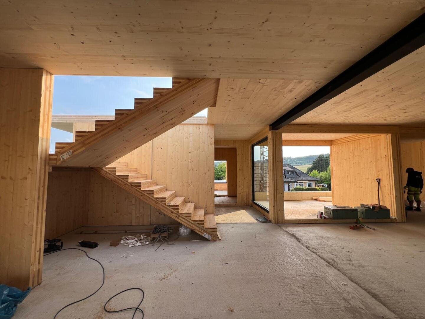 Interior of a modern house under construction, featuring unfinished wooden walls, open spaces, a wooden staircase, exposed wiring, and tools on the floor. Large windows offer views of greenery and neighboring houses outside.
