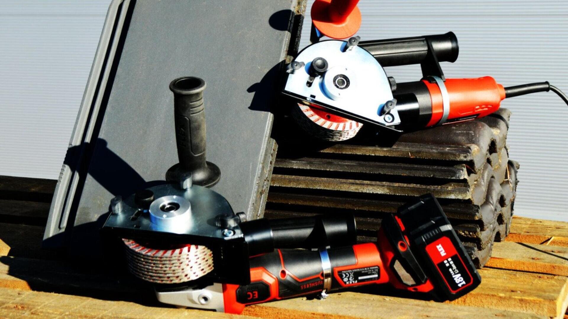 Two red and black angle grinders rest on stacked metal rods and a gray board, on a wooden pallet. The scene appears to be set outdoors in bright sunlight.