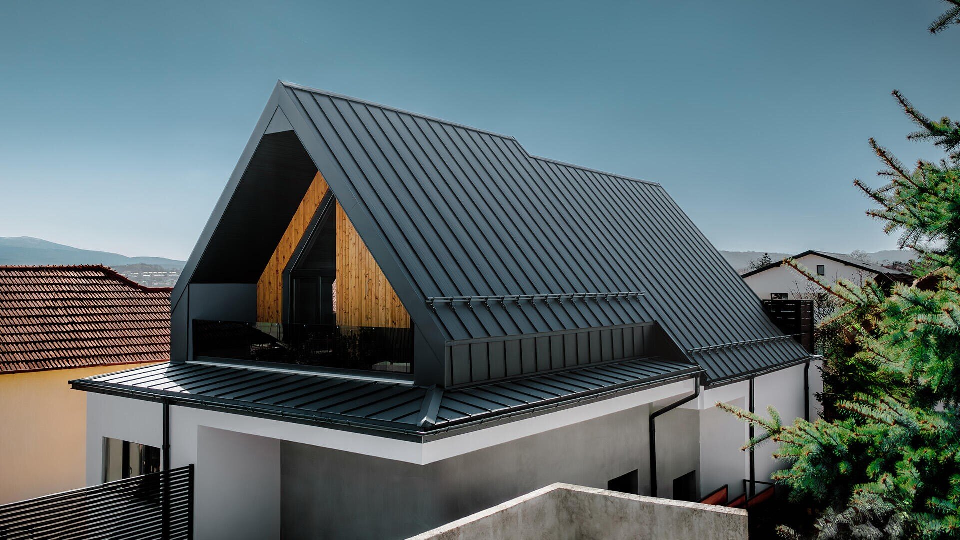 Modernes Haus mit steilem, dunklem Metallgiebeldach und großem Dreiecksfenster, mit Holzakzenten im Obergeschoss. Das Haus ist von anderen Häusern und Grünflächen unter einem klaren blauen Himmel umgeben.
