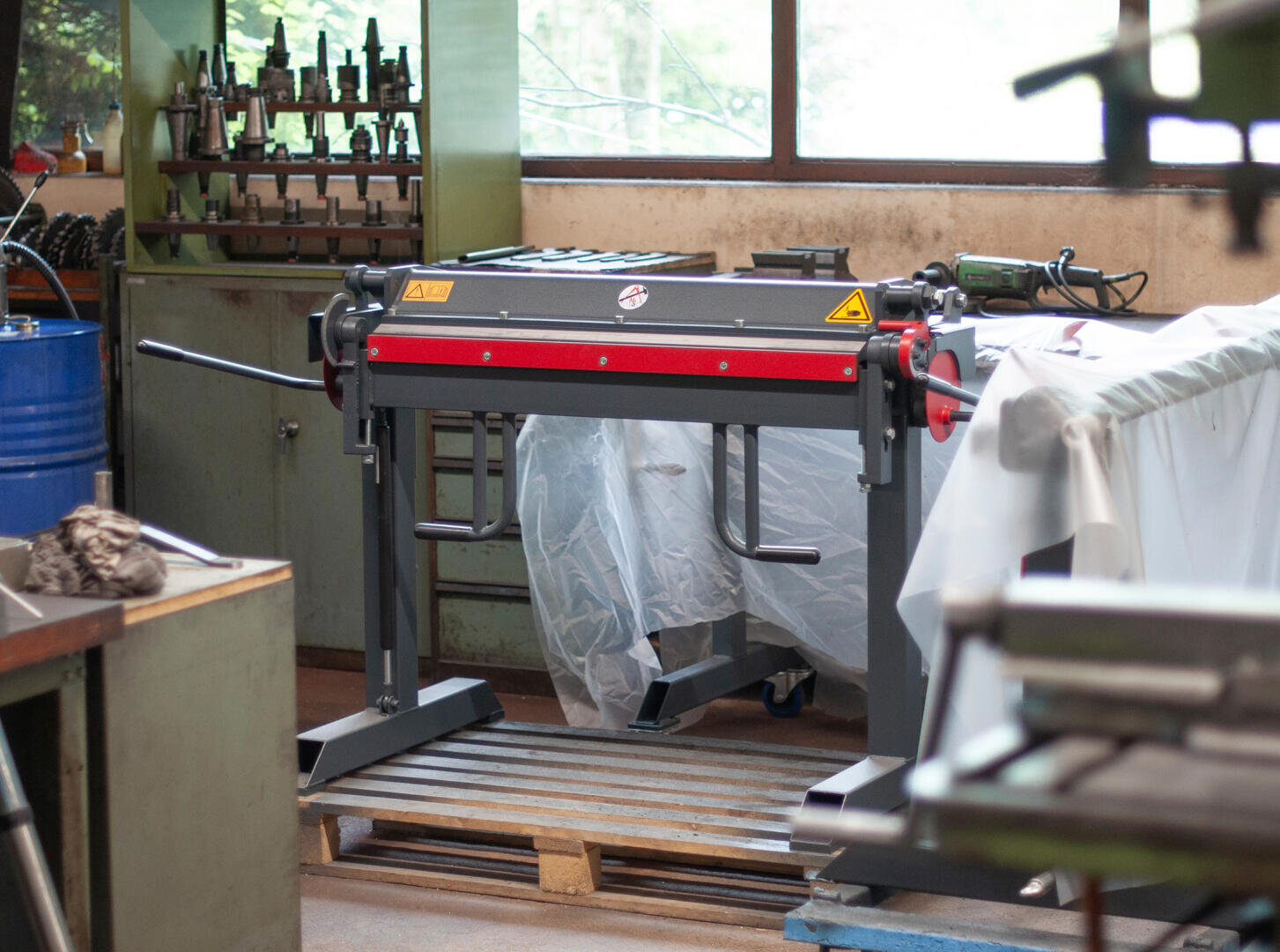 Eine Metallbiegemaschine steht in einer Werkstatt, die von Werkzeugen, Maschinen und abgedeckten Geräten umgeben ist und durch die großen Fenster natürliches Licht einfällt.