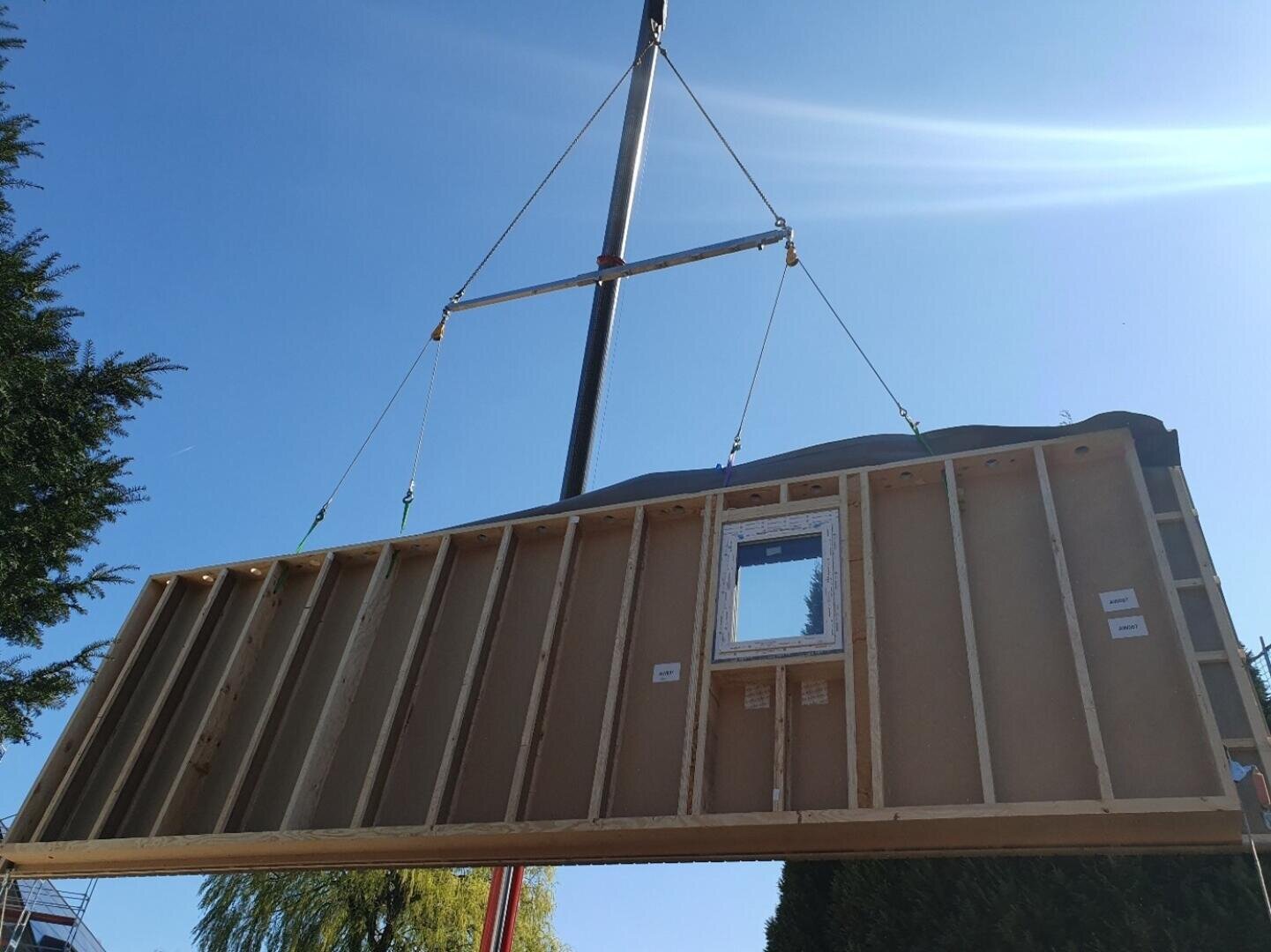A large, prefabricated wooden wall with a window opening is lifted by a crane against a bright blue sky, probably for installation in a building.