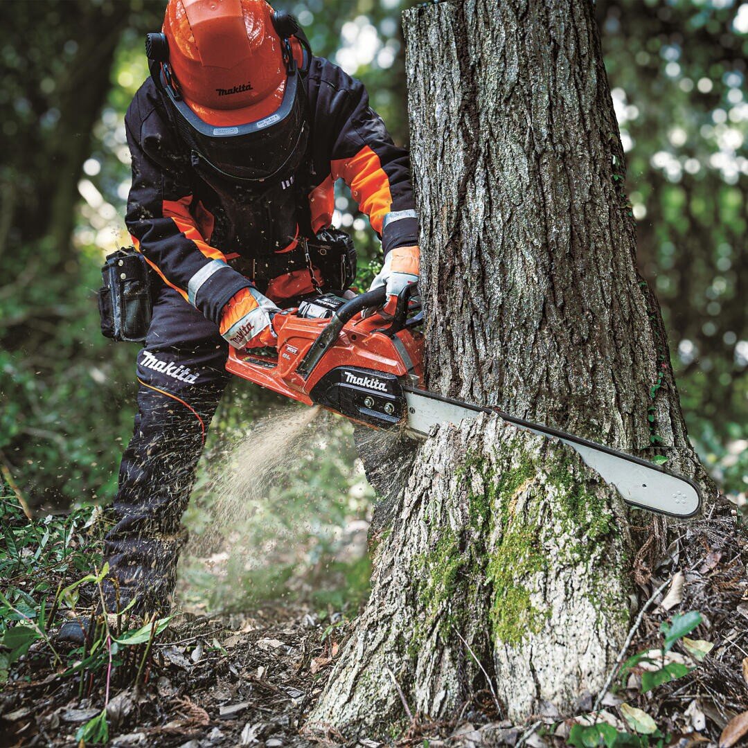 Eine Person, die Schutzkleidung und einen Helm trägt, fällte mit einer Makita-Kettensäge einen Baum in einem Wald, wobei Holzspäne umherflogen.