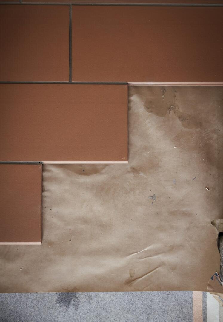 Ein Abschnitt aus rötlich-braunen rechteckigen Fliesen ist teilweise über einem Blatt zerknitterten braunen Papiers verlegt, das einen Teil einer grauen Fläche darunter bedeckt. Die Fliesen sind in einem gestaffelten Ziegelmuster angeordnet.