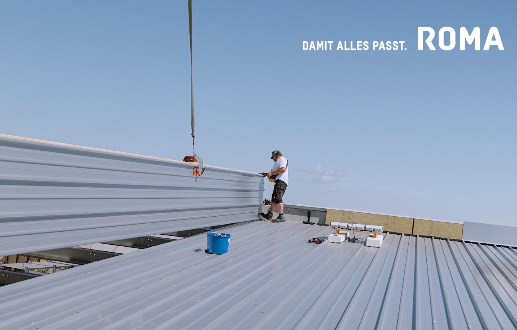 Ein Arbeiter in kurzen Hosen und mit Mütze steht auf einem Metalldach und montiert unter blauem Himmel große Metallplatten mit einem Kran. Auf dem Dach befinden sich Werkzeuge und ein blauer Eimer. DAMIT ALLES PASST. In der Ecke erscheint der Schriftzug ROMA.