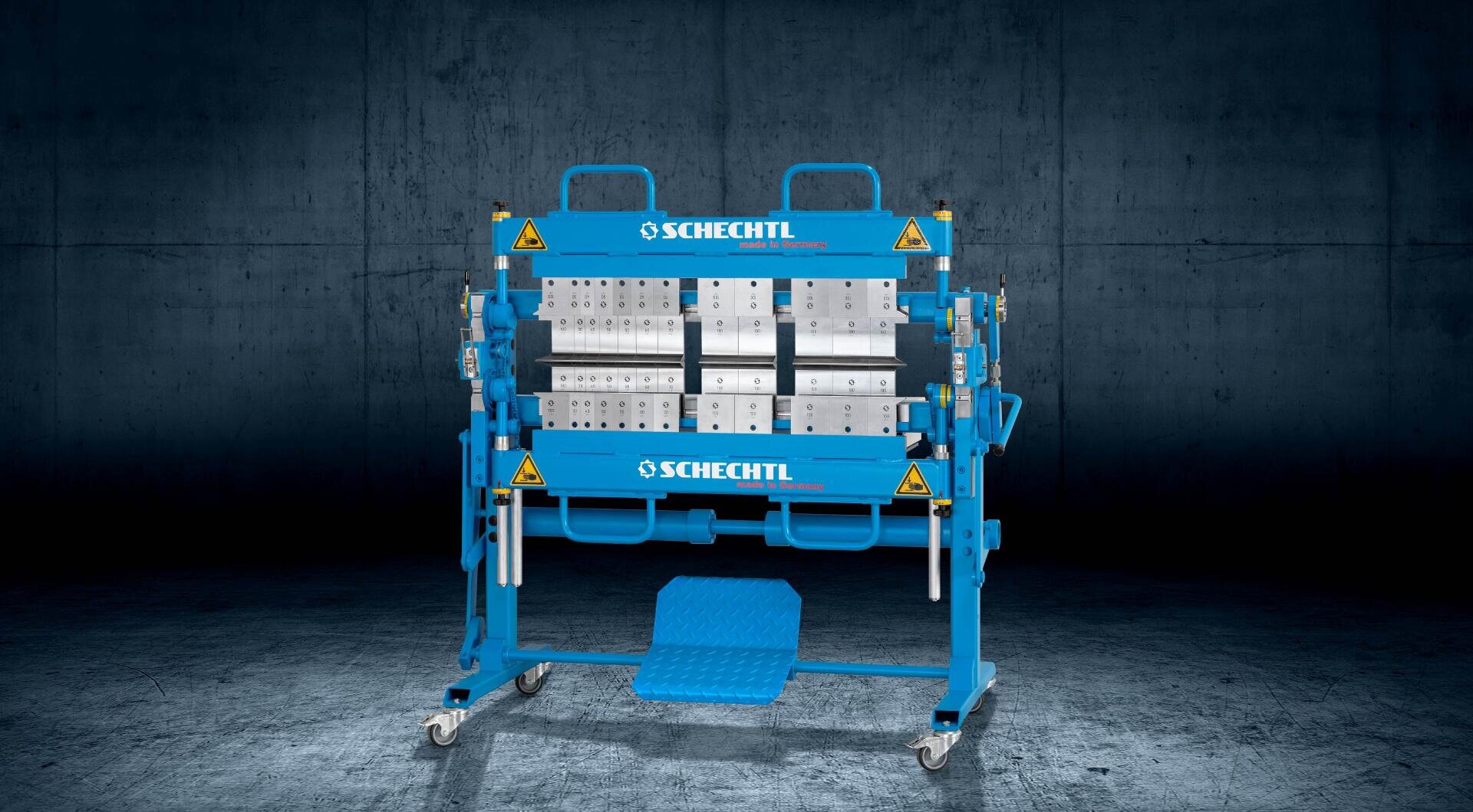 Eine blaue Schechtl-Metallbearbeitungsmaschine mit sichtbaren Rollen und Hebeln steht auf einem Radgestell vor einem dunklen, industriellen Hintergrund. An der Maschine sind gelbe Warnaufkleber zu sehen.