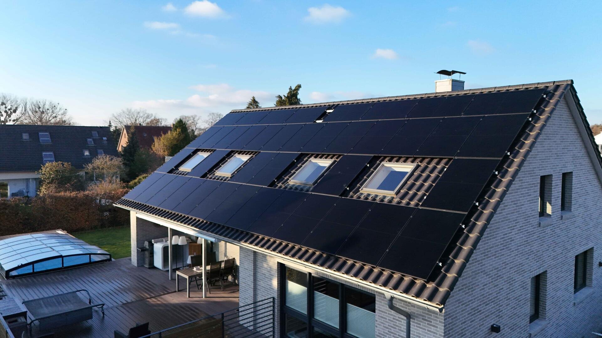 A modern house with a pitched roof covered with black solar panels. There are several skylights on the roof and the house is surrounded by a garden with patio furniture. The sky is mostly clear with a few clouds.