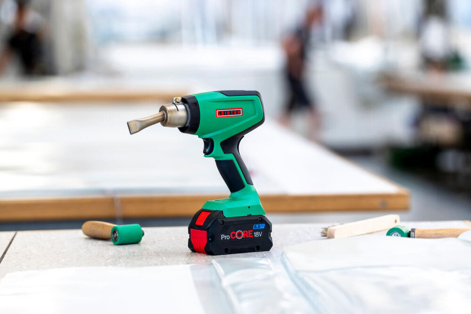 A green and black cordless heat gun with digital display stands upright on a worktable surrounded by plastic panels and hand tools in a bright workshop environment.
