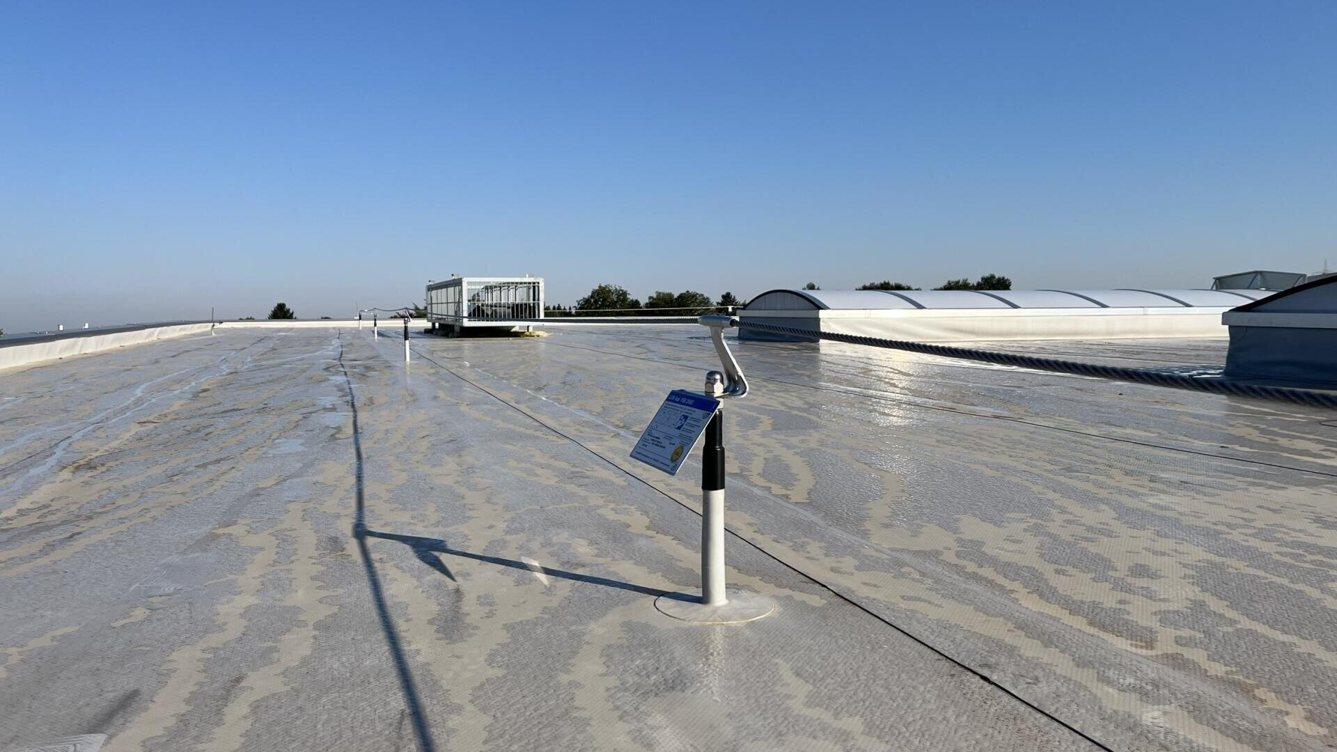 A flat, sunlit rooftop with light-colored waterproofing, safety line posts, and a warning sign in the foreground. Skylights, vents, and a clear blue sky are visible in the background.