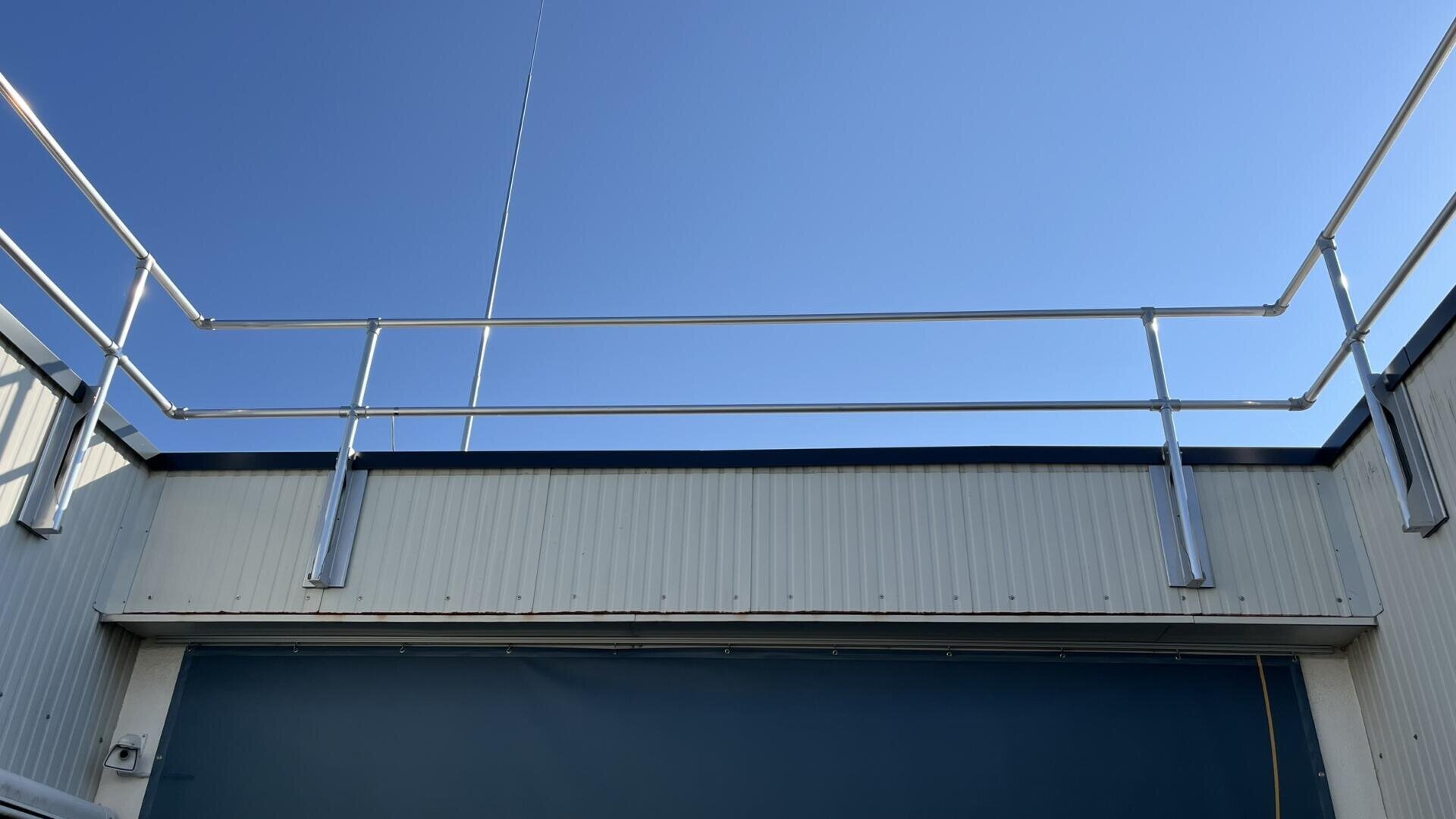 A rooftop with metal safety railings against a clear blue sky. The building has a corrugated exterior wall and a blue panel beneath the rooftop.