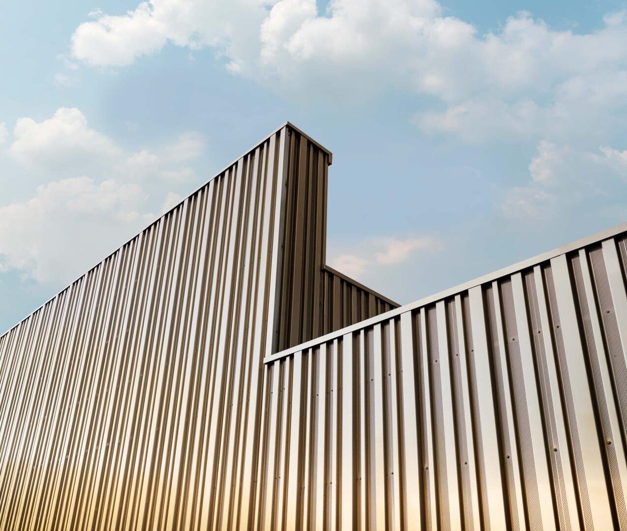 Moderne Gebäudefassade aus Wellblech mit klaren Linien und scharfen Winkeln vor einem blauen Himmel mit weichen Wolken. Das Sonnenlicht wirft subtile Reflexionen auf die metallische Oberfläche.