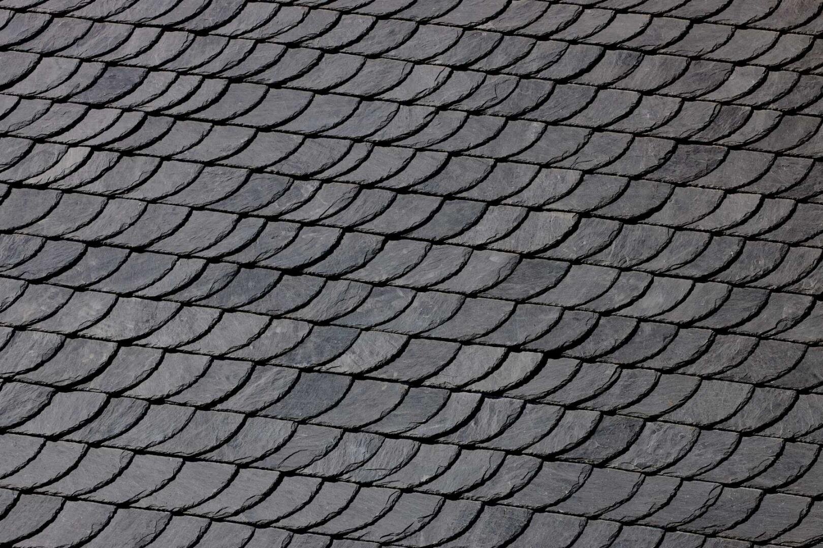 Nahaufnahme von sich überlappenden dunkelgrauen Schieferdachziegeln, die in sauberen, diagonalen Reihen angeordnet sind und eine strukturierte und gemusterte Oberfläche bilden.